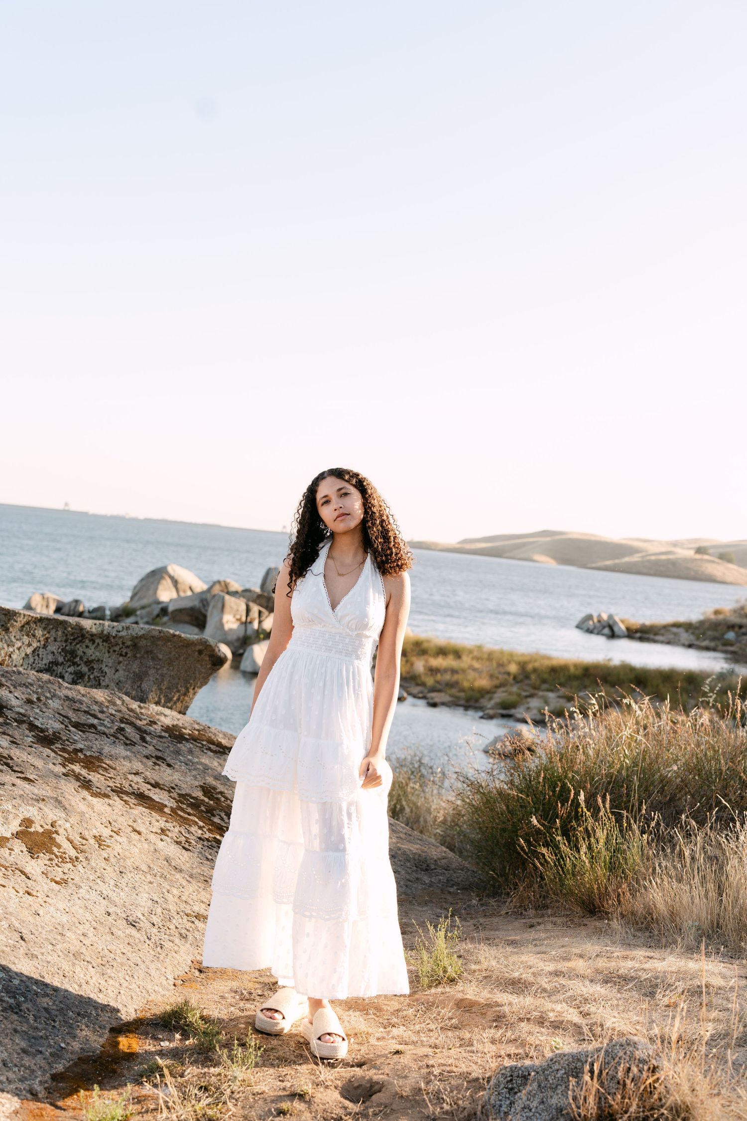 Young woman in white maxi dress standing on rocky coastal landscape with ocean and sandy hills in background.