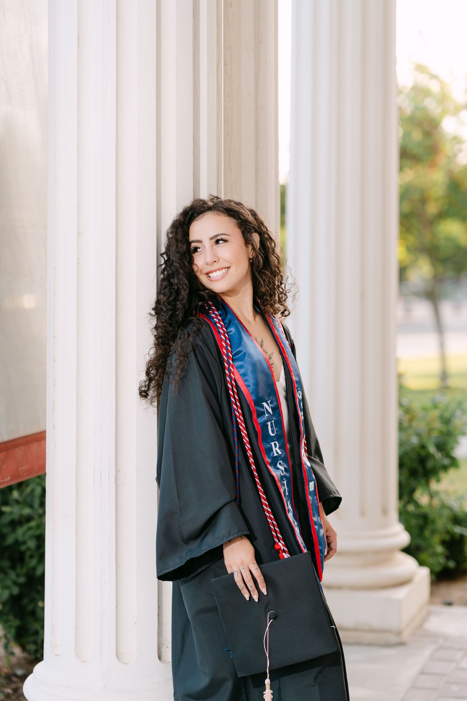 Smiling nursing graduate in black gown with red cords and nursing stole poses between white columns outdoors.