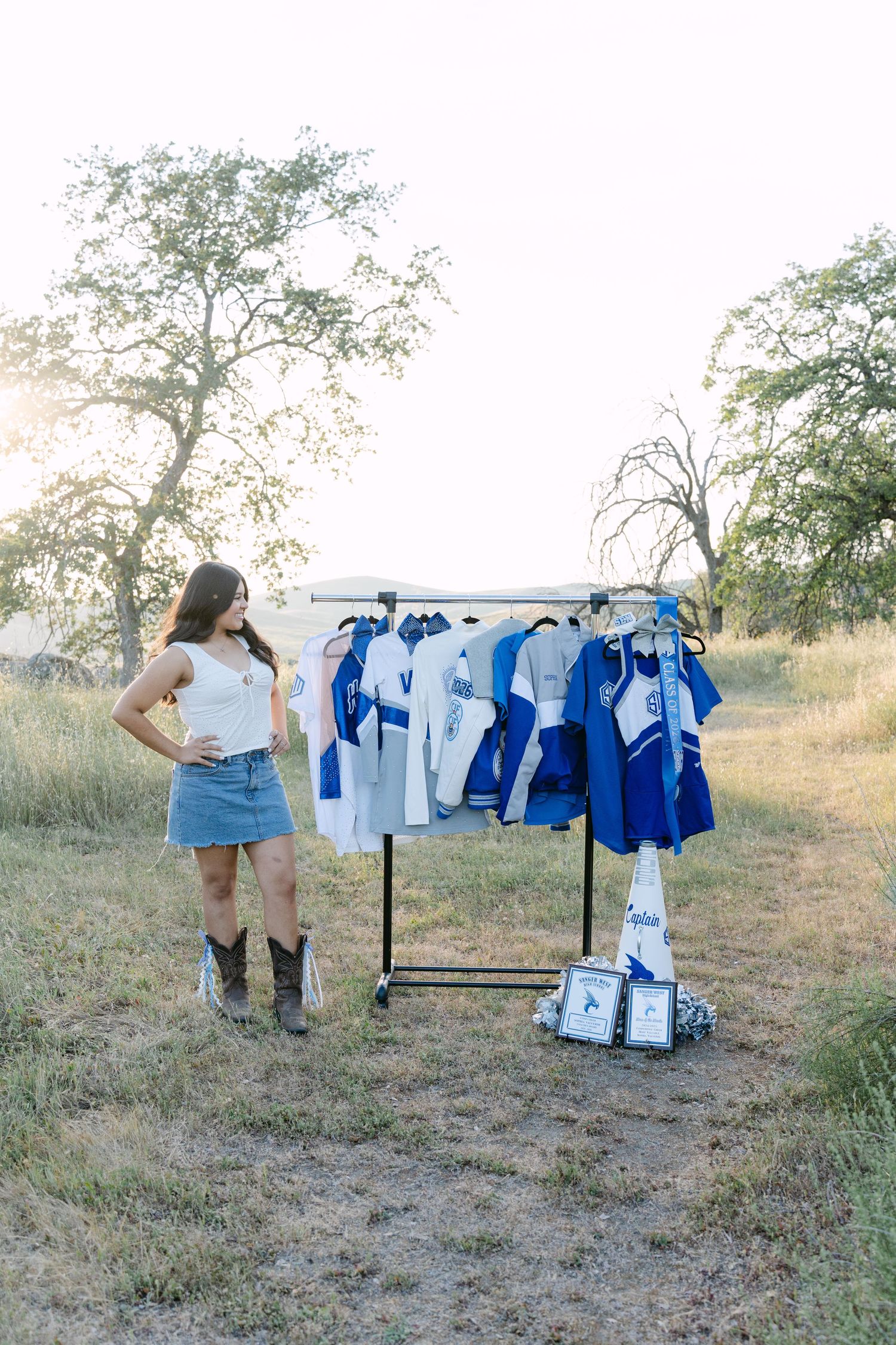Young woman in denim skirt and cowboy boots stands beside outdoor clothing rack displaying blue and white jerseys.