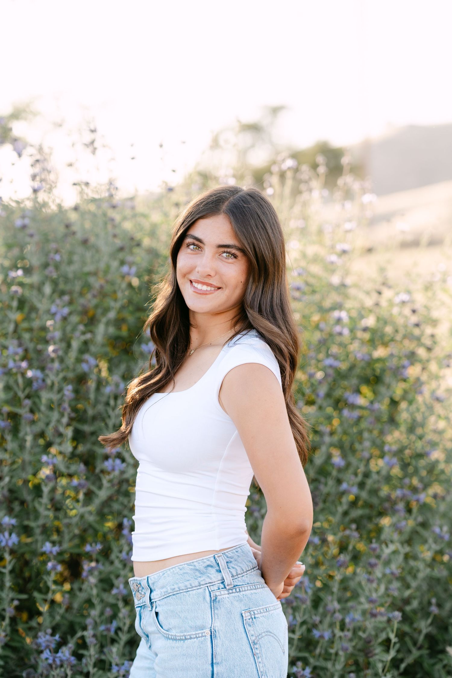 Young woman in white crop top and jeans smiling outdoors among purple wildflowers in golden sunlight.