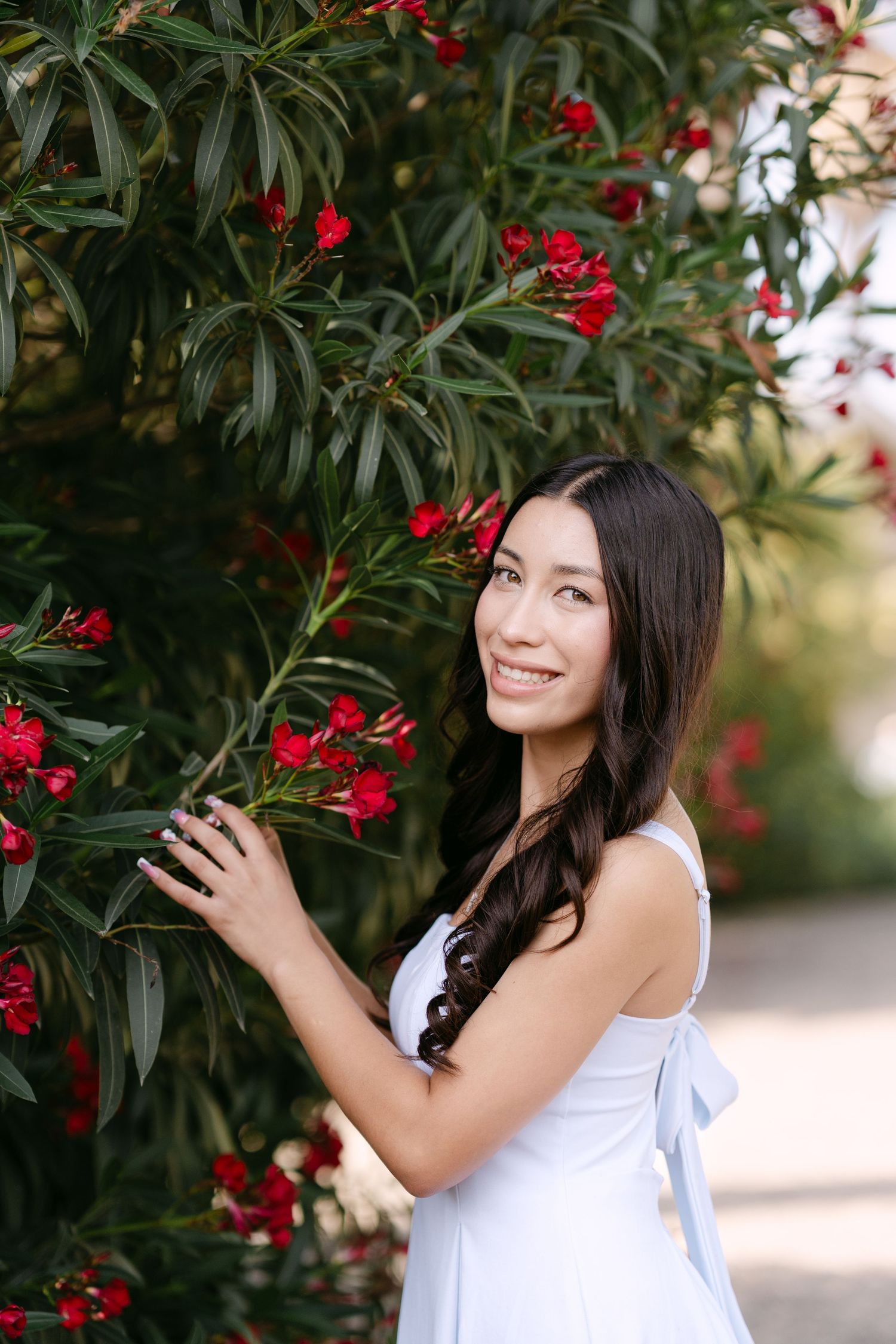 Young woman in white dress smiling beside red oleander flowers in a lush green garden.