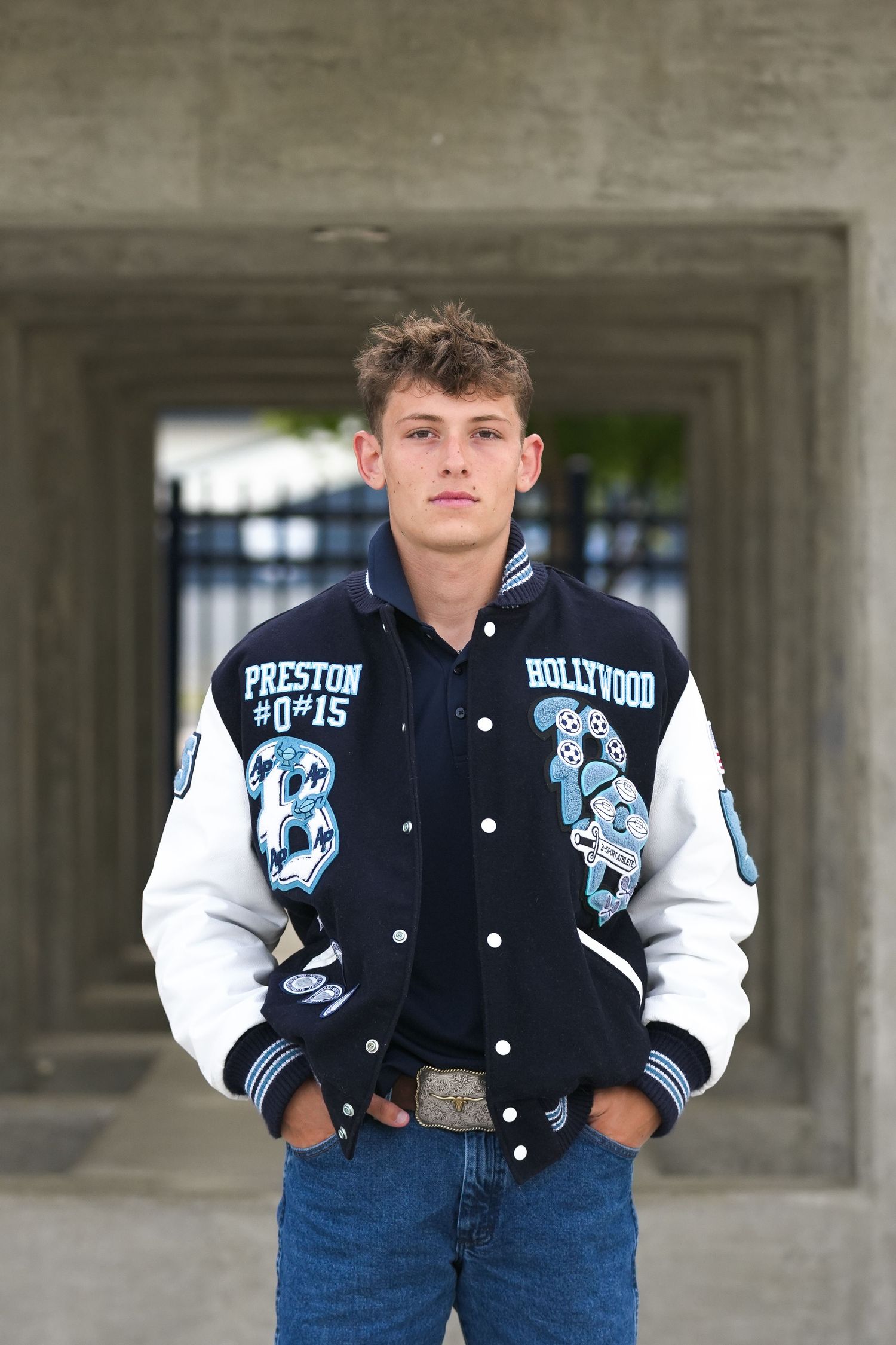 Student wearing varsity letterman jacket poses against concrete building backdrop.