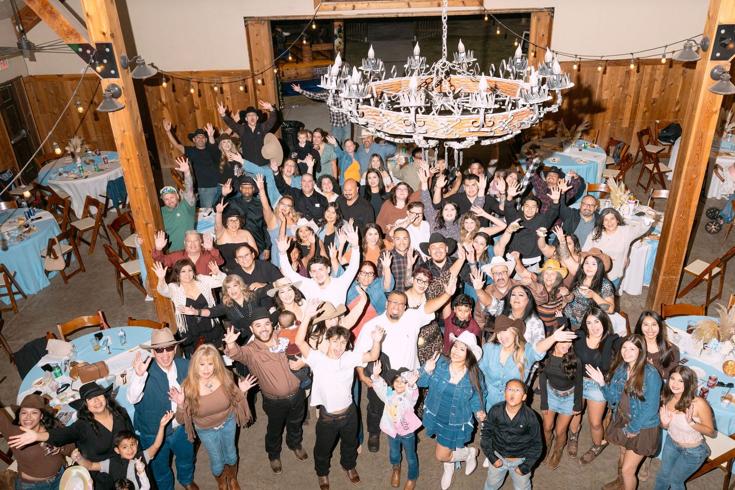 Large crowd of wedding guests dancing and celebrating together on a wooden dance floor at an indoor reception venue.