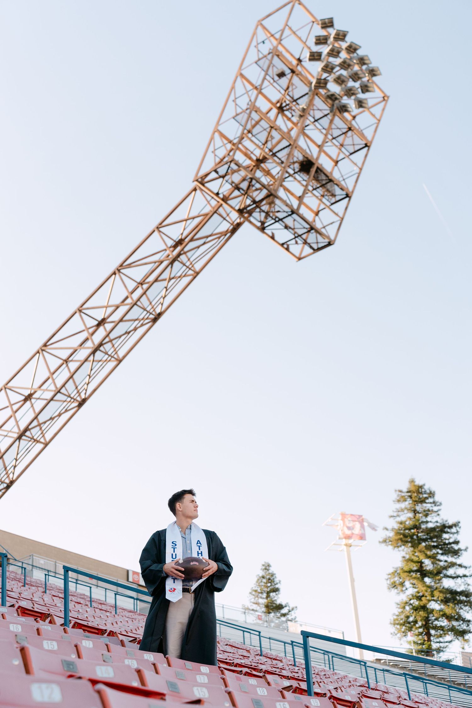 Graduate in cap and gown stands in stadium bleachers beneath towering floodlight against clear sky.