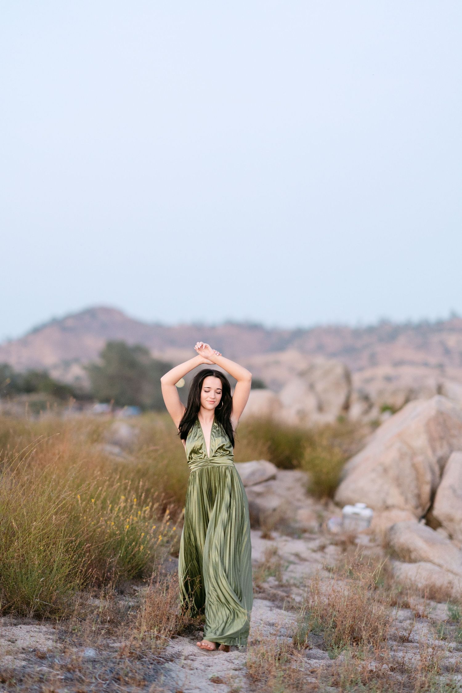 Woman in flowing green dress with arms raised in rocky desert landscape.