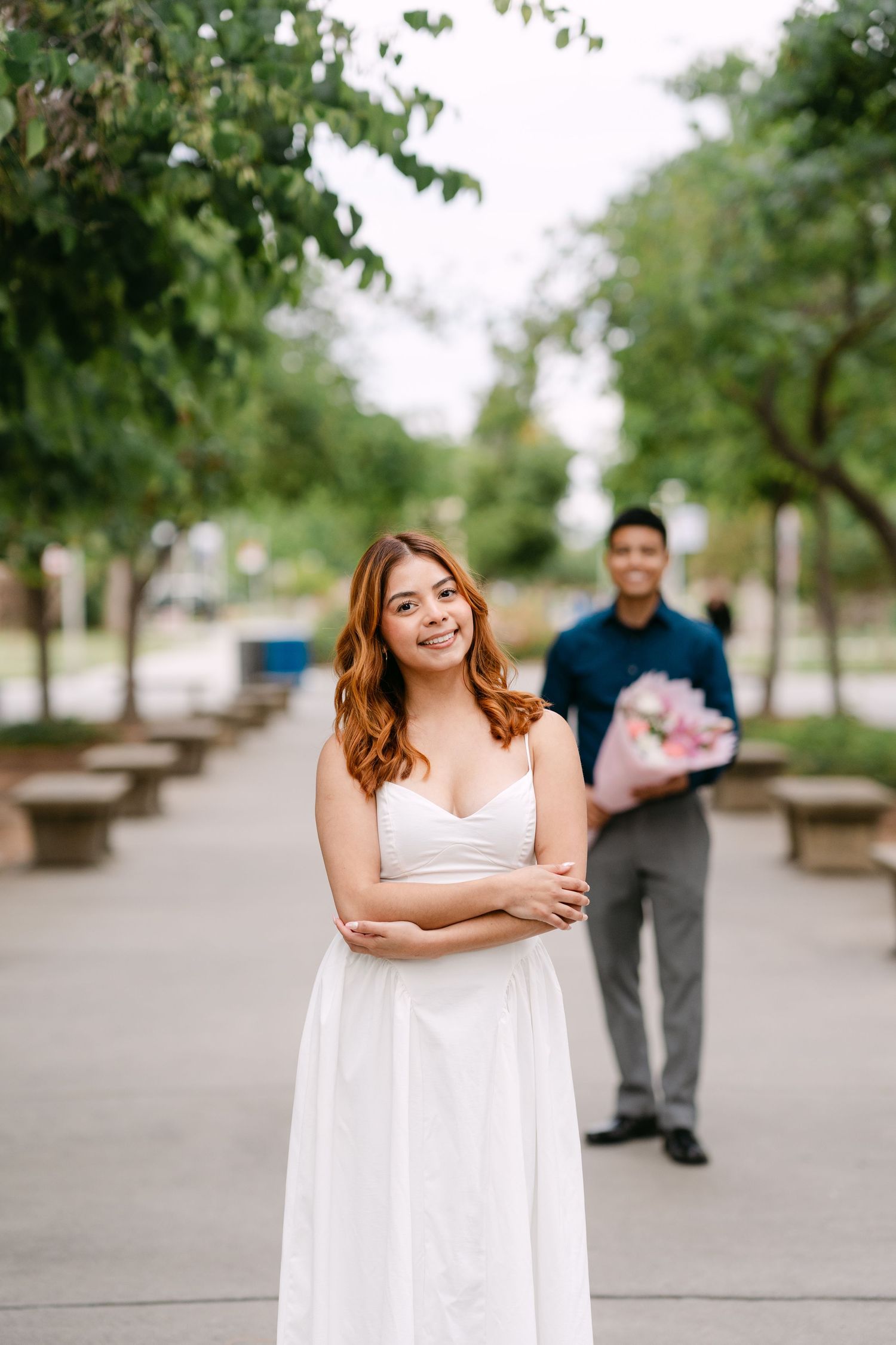 Young woman in white dress stands with arms crossed outdoors as man holds pink flowers behind her.