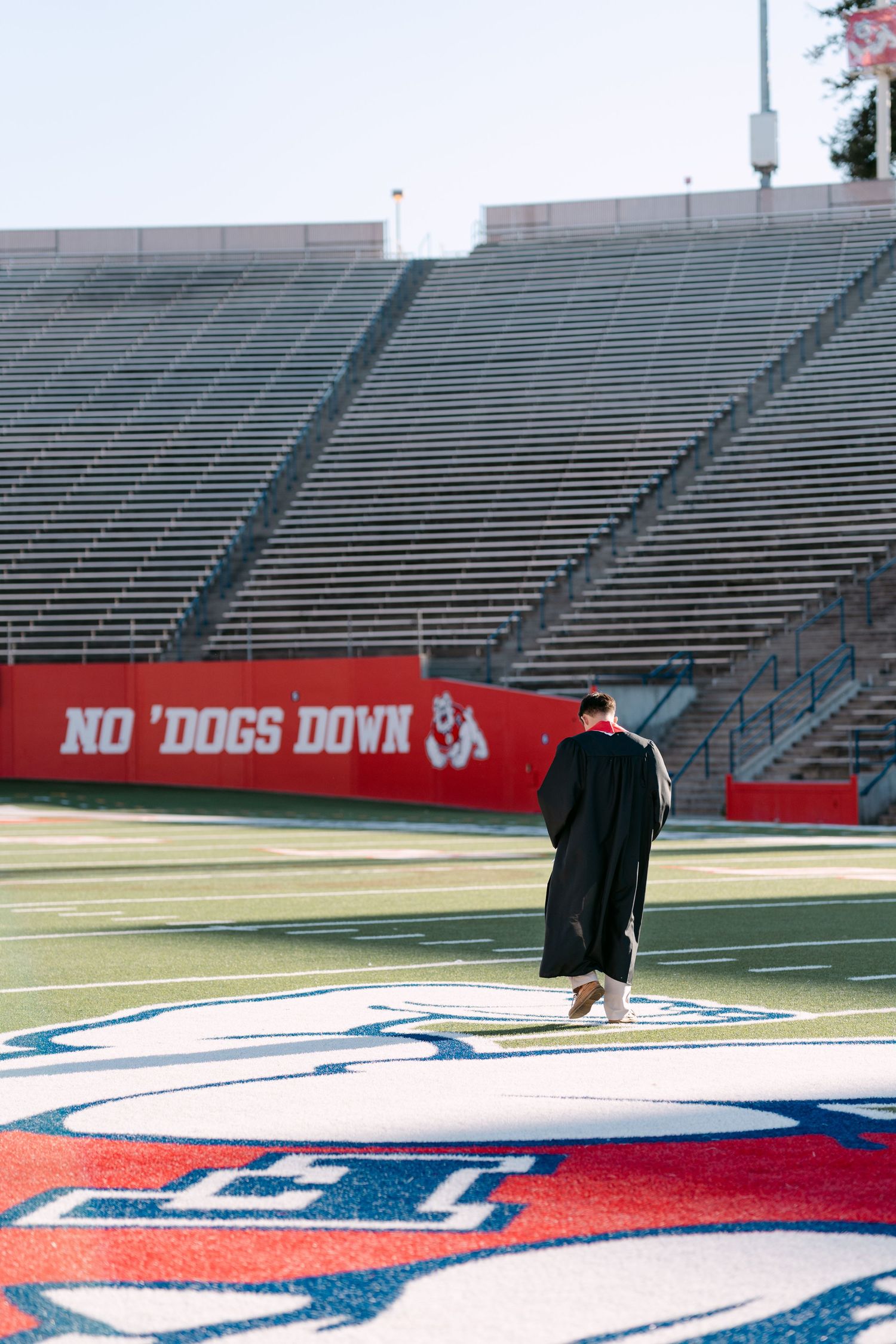 Graduate in black cap and gown stands on Fresno State Bulldogs football field logo at empty Bulldog Stadium.