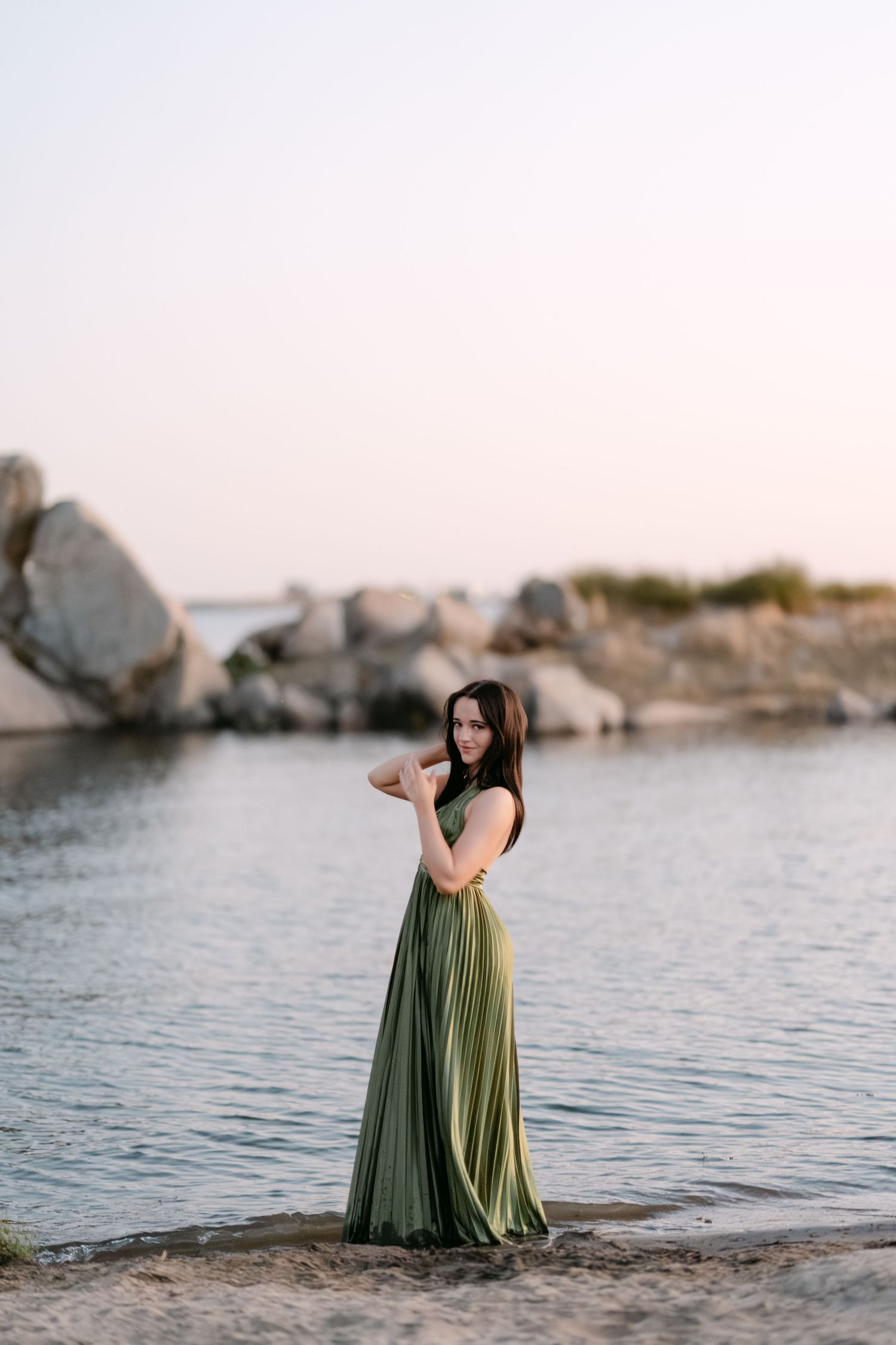 Woman in elegant green maxi dress posing on rocky beach at sunset.