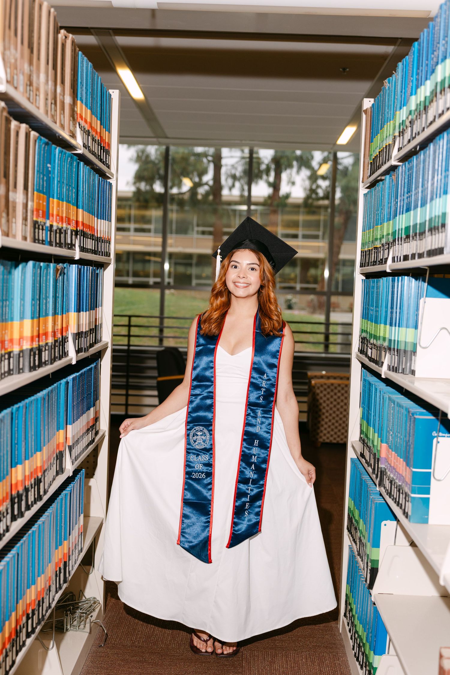 Smiling graduate in white gown and cap poses between library bookshelves wearing a blue honor stole.