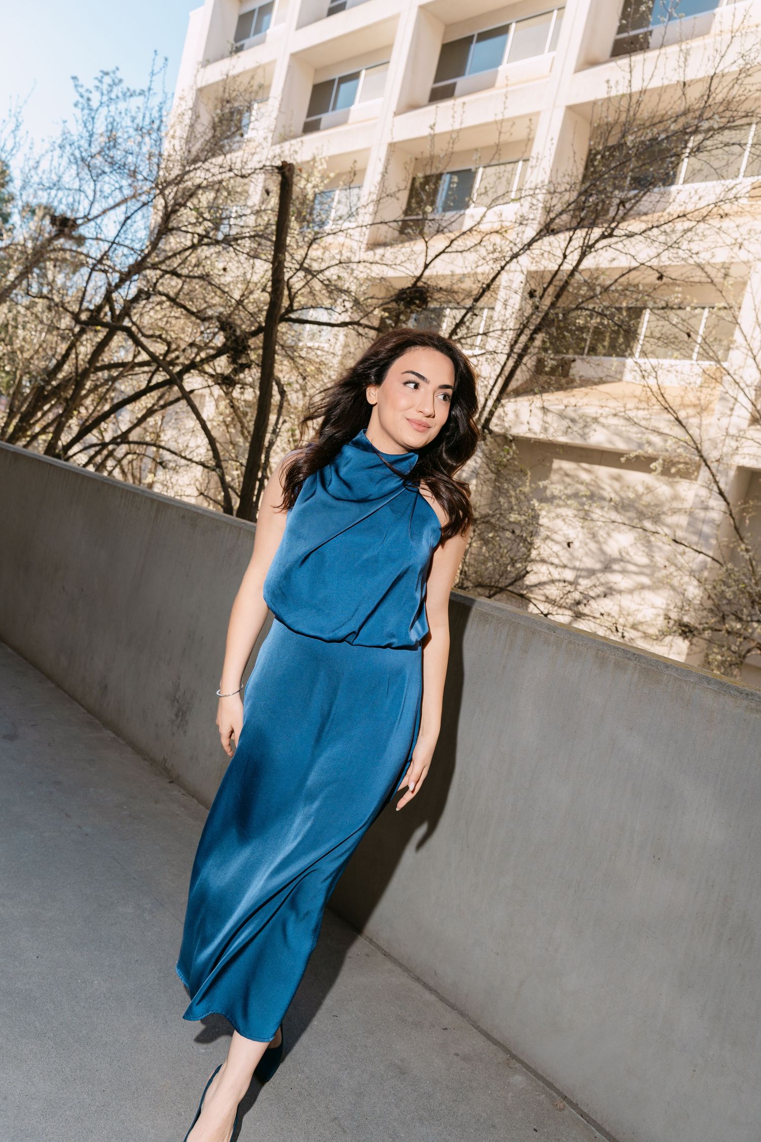 Woman in flowing blue maxi dress poses against concrete wall with apartment building in background.
