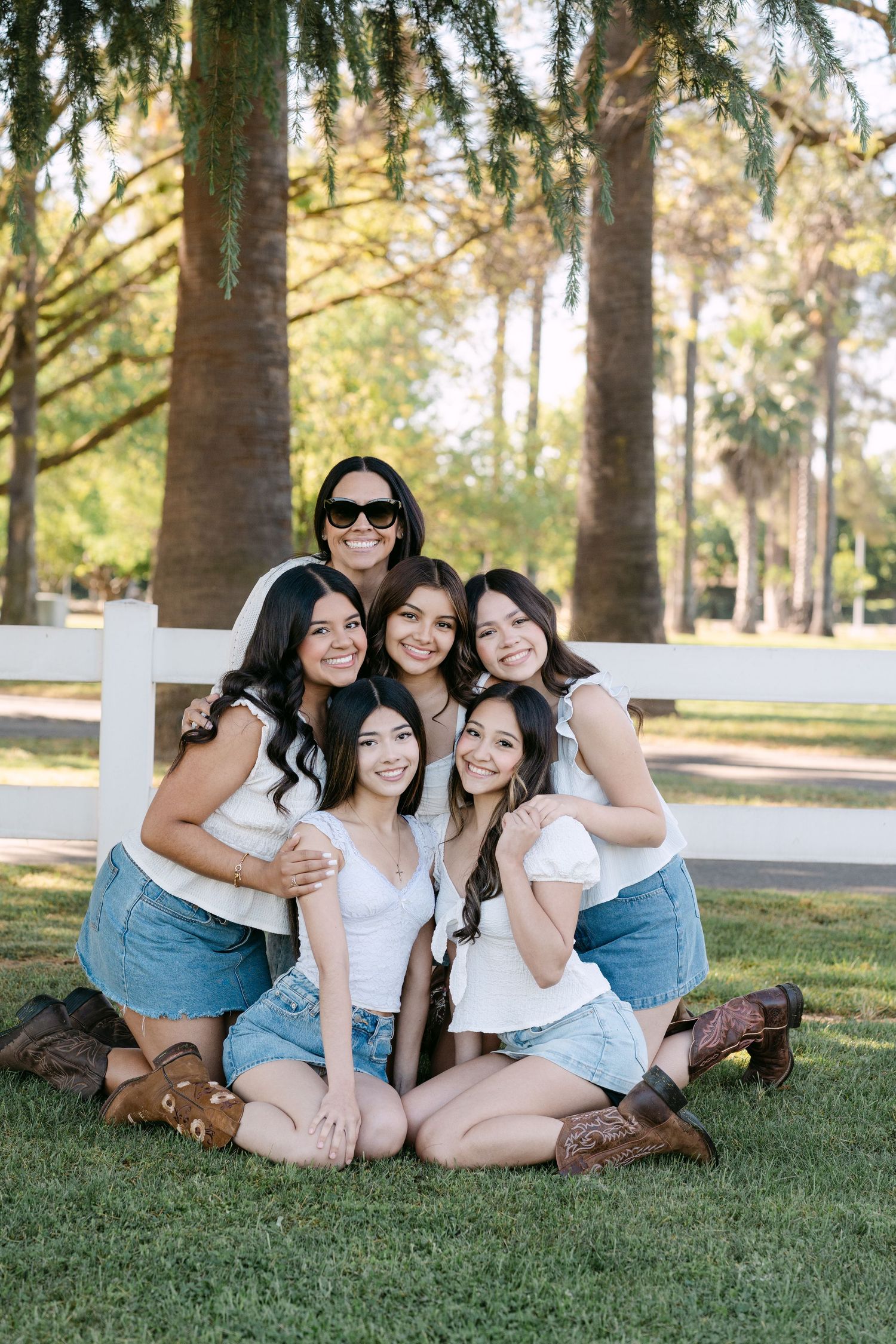 Six young women pose together on green grass in a park, wearing matching white tops and denim skirts with cowboy boots.