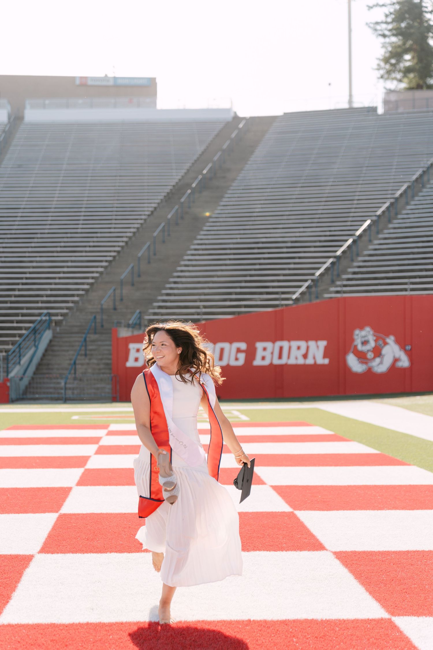Graduate in white dress and red stole walks on Fresno State Bulldog Born stadium field during graduation photos.
