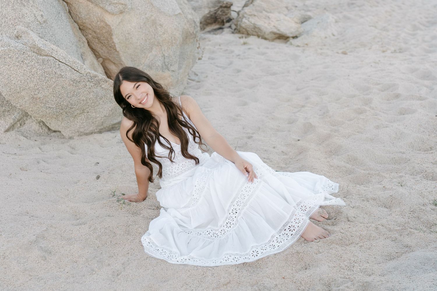 Young woman in white lace dress sitting on sandy beach, smiling, with large rocks behind her.