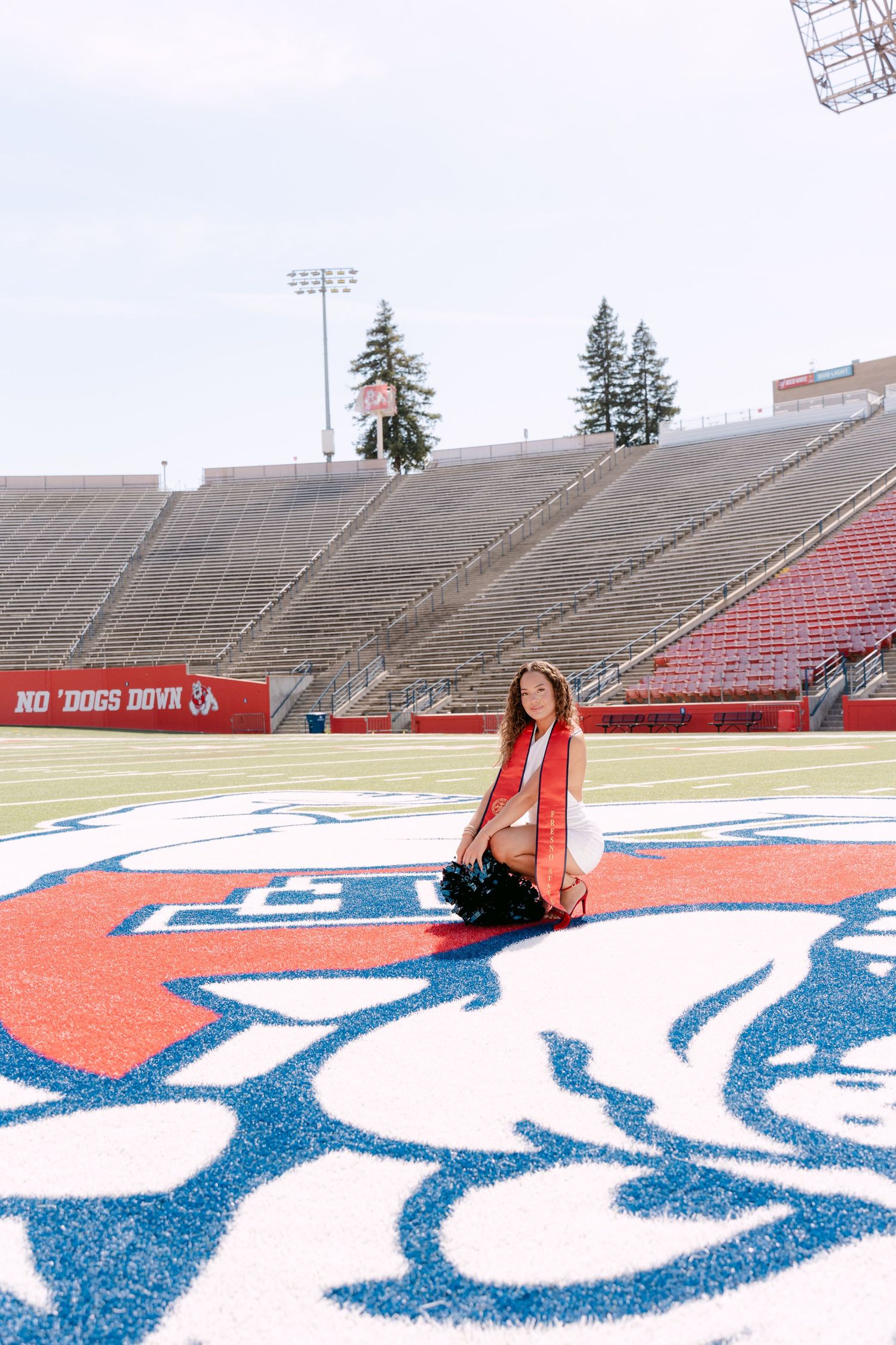 Graduate poses on Fresno State Bulldogs logo at Bulldog Stadium wearing red sash, with empty bleachers behind her.