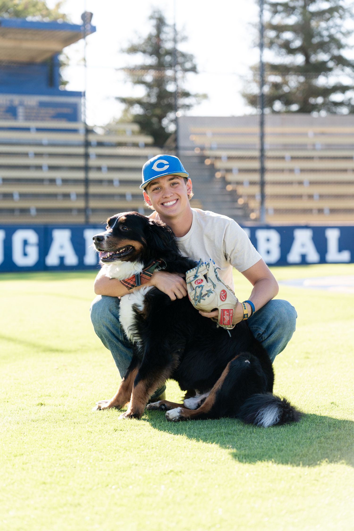 Baseball player poses with friendly black and brown dog on sunny sports field with stadium bleachers in background.