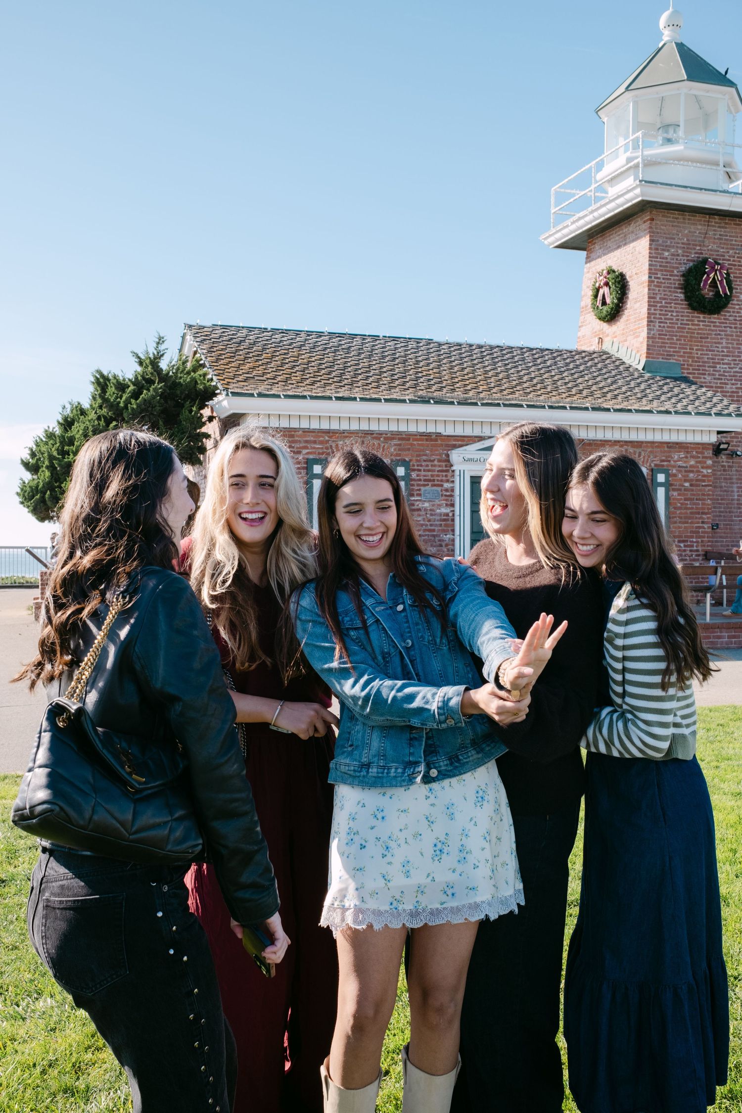 A group of friends laugh together while taking a selfie outside a lighthouse with brick walls on a sunny day.