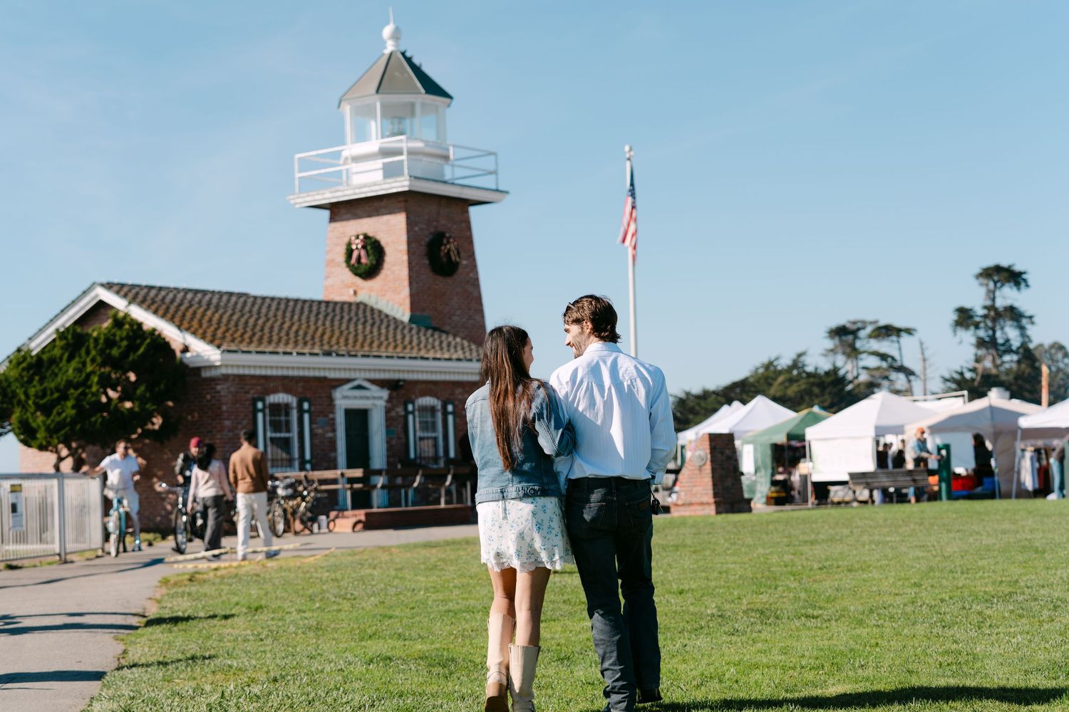 A couple walks on a grassy lawn near a lighthouse-style building with an American flag on a sunny day.