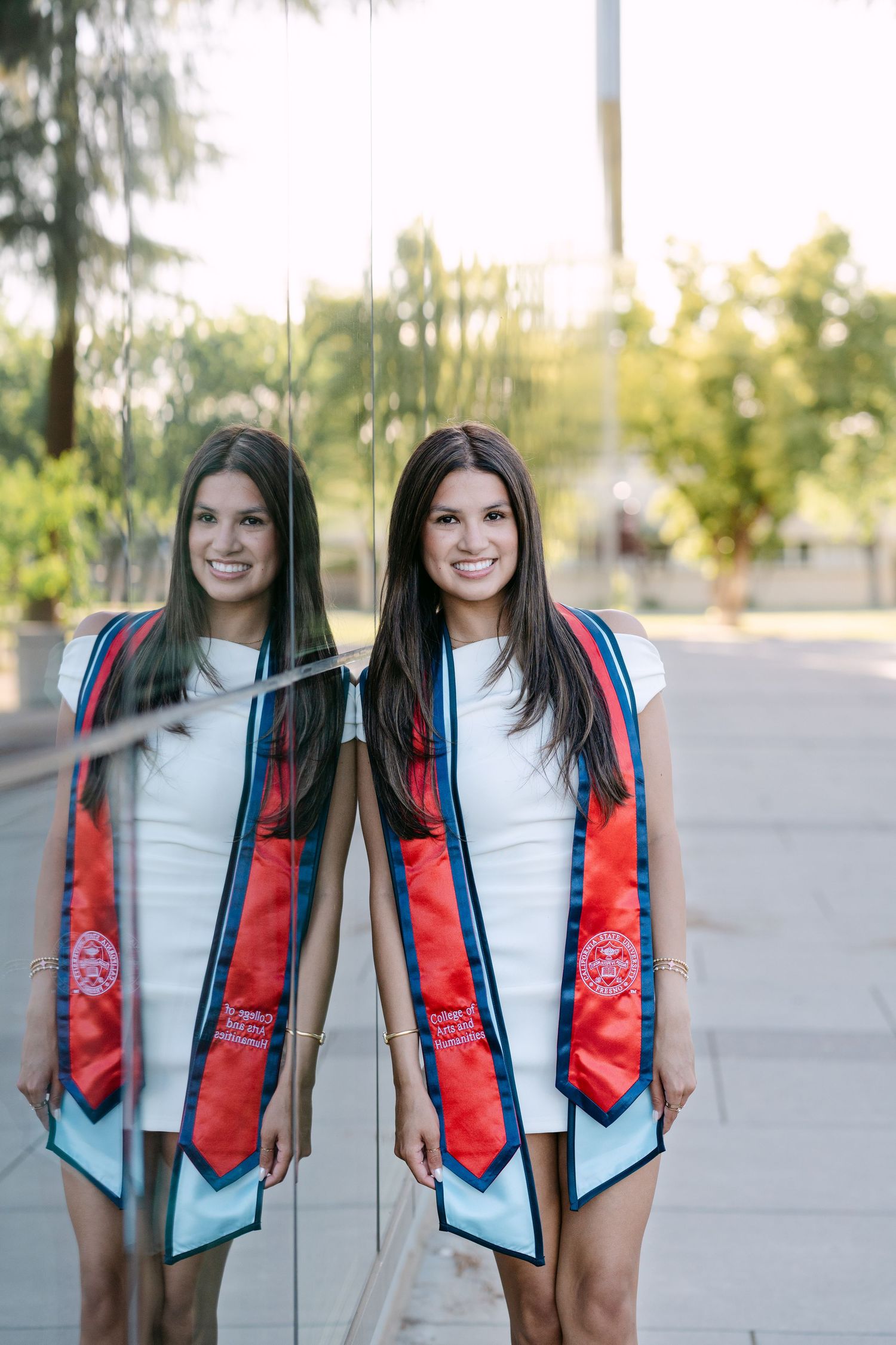 Young woman in white dress and red graduation stole smiles beside her reflection in glass outdoors.