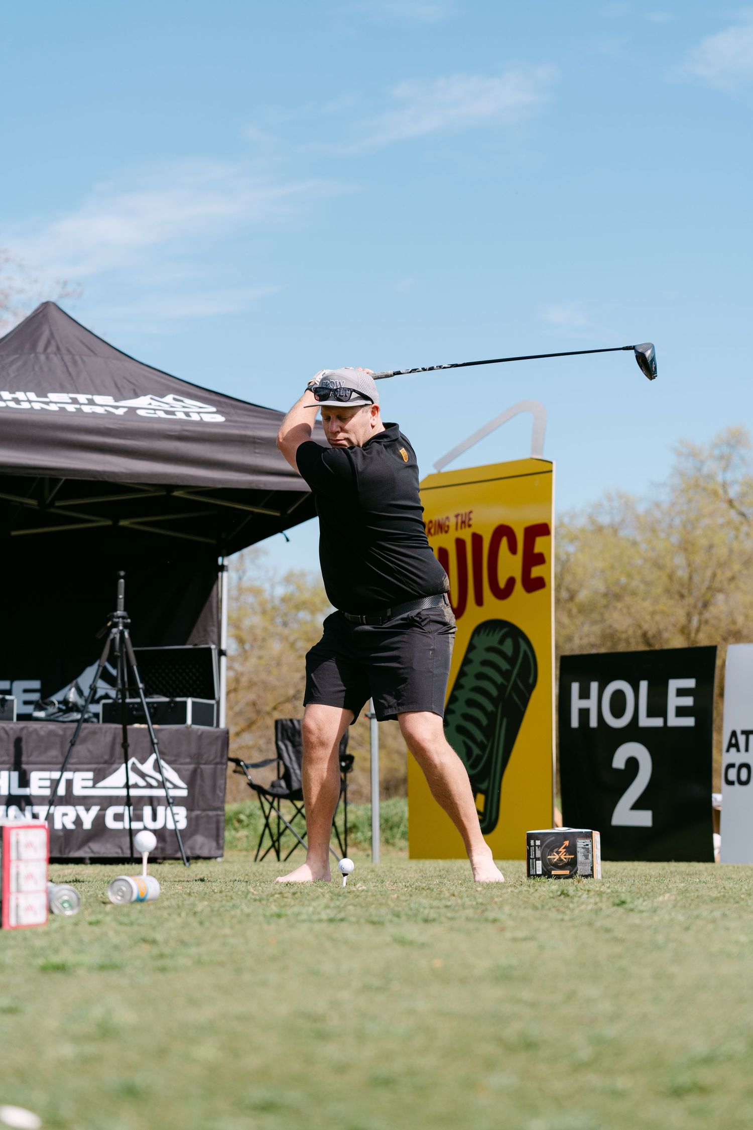Golfer swinging a club at hole 2 during an Athlete Country Club golf tournament on a sunny day.