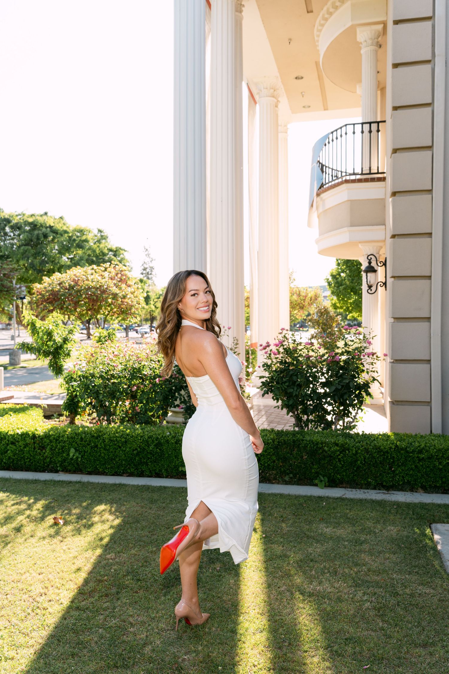 Woman in white dress holding red heels, smiling outdoors near elegant building with columns and balcony.
