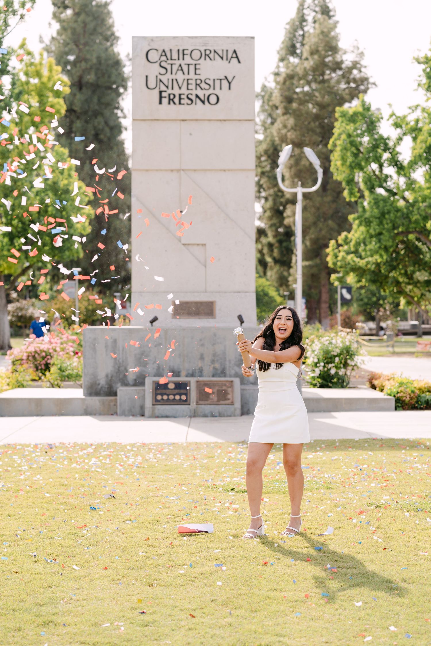 Joyful graduate celebrates with confetti at California State University Fresno sign in white dress.