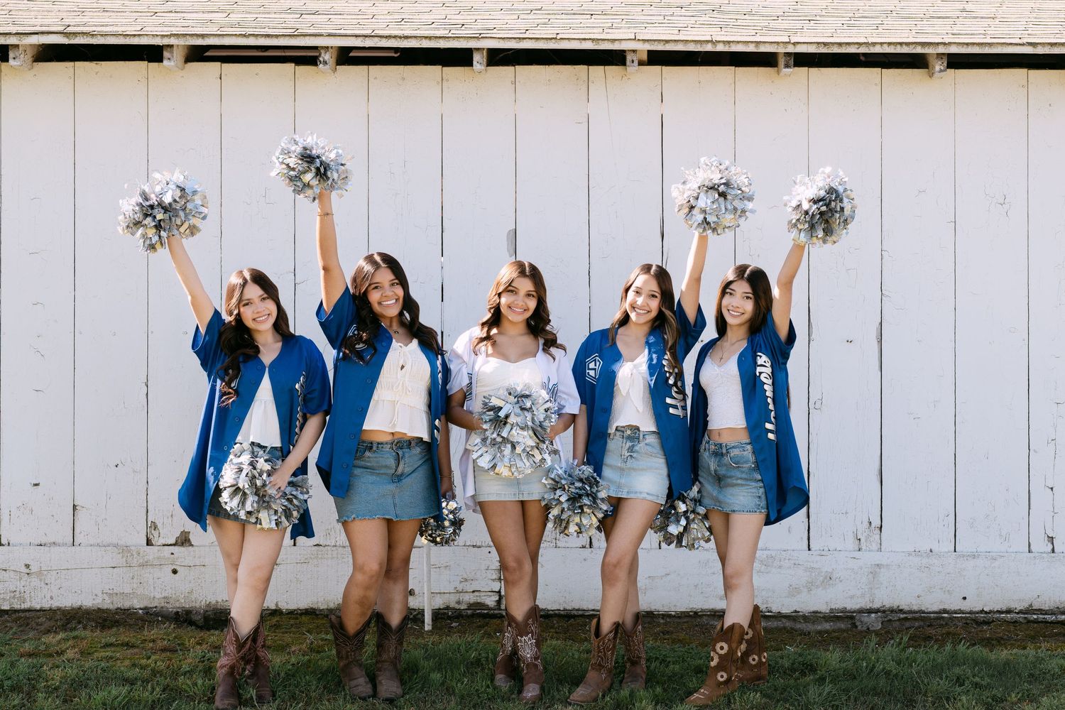 Five cheerleaders in blue robes and denim skirts pose with silver pom-poms in front of a white barn wall.