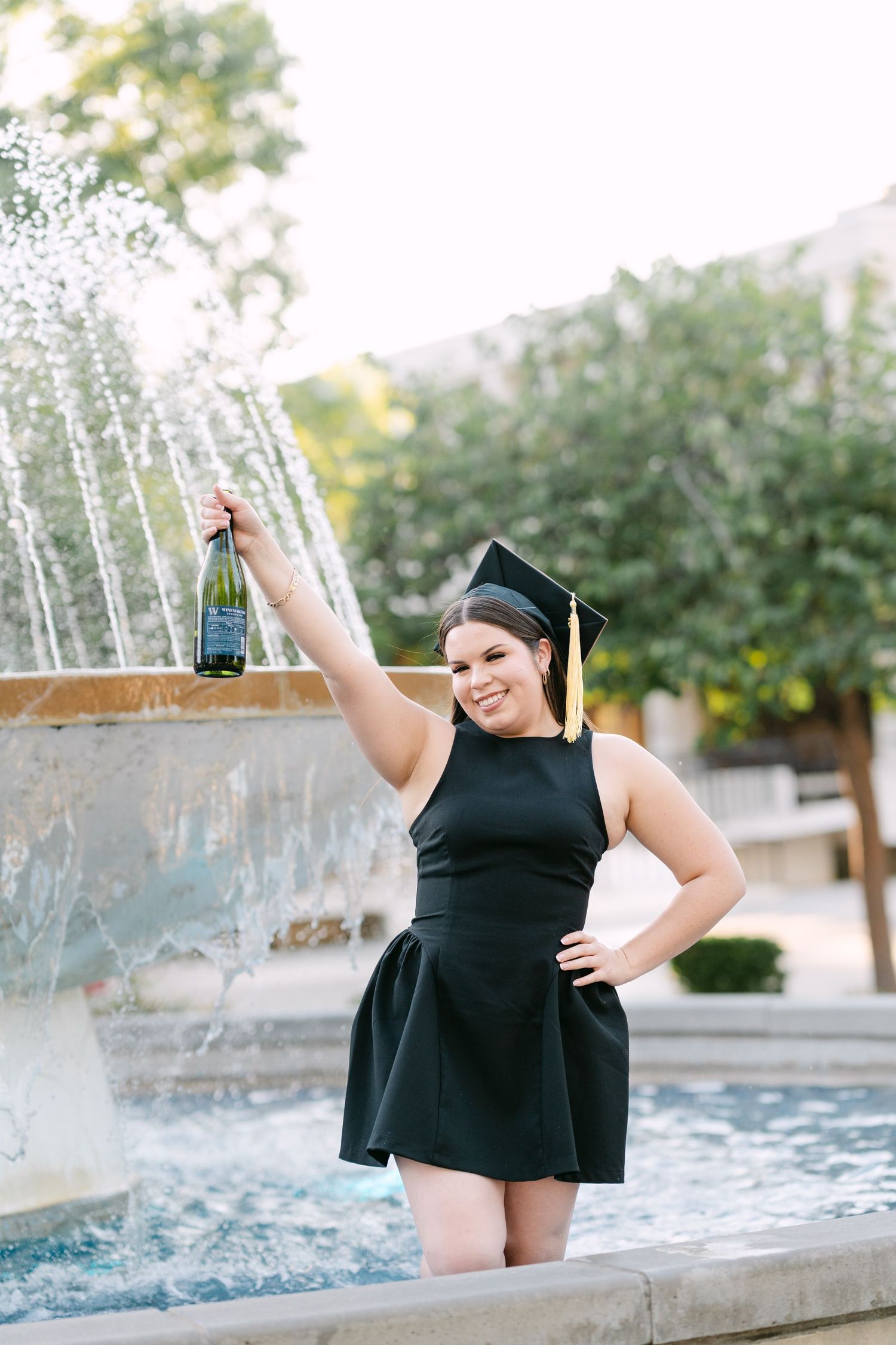 Graduate in black dress and cap holding champagne bottle by campus fountain.