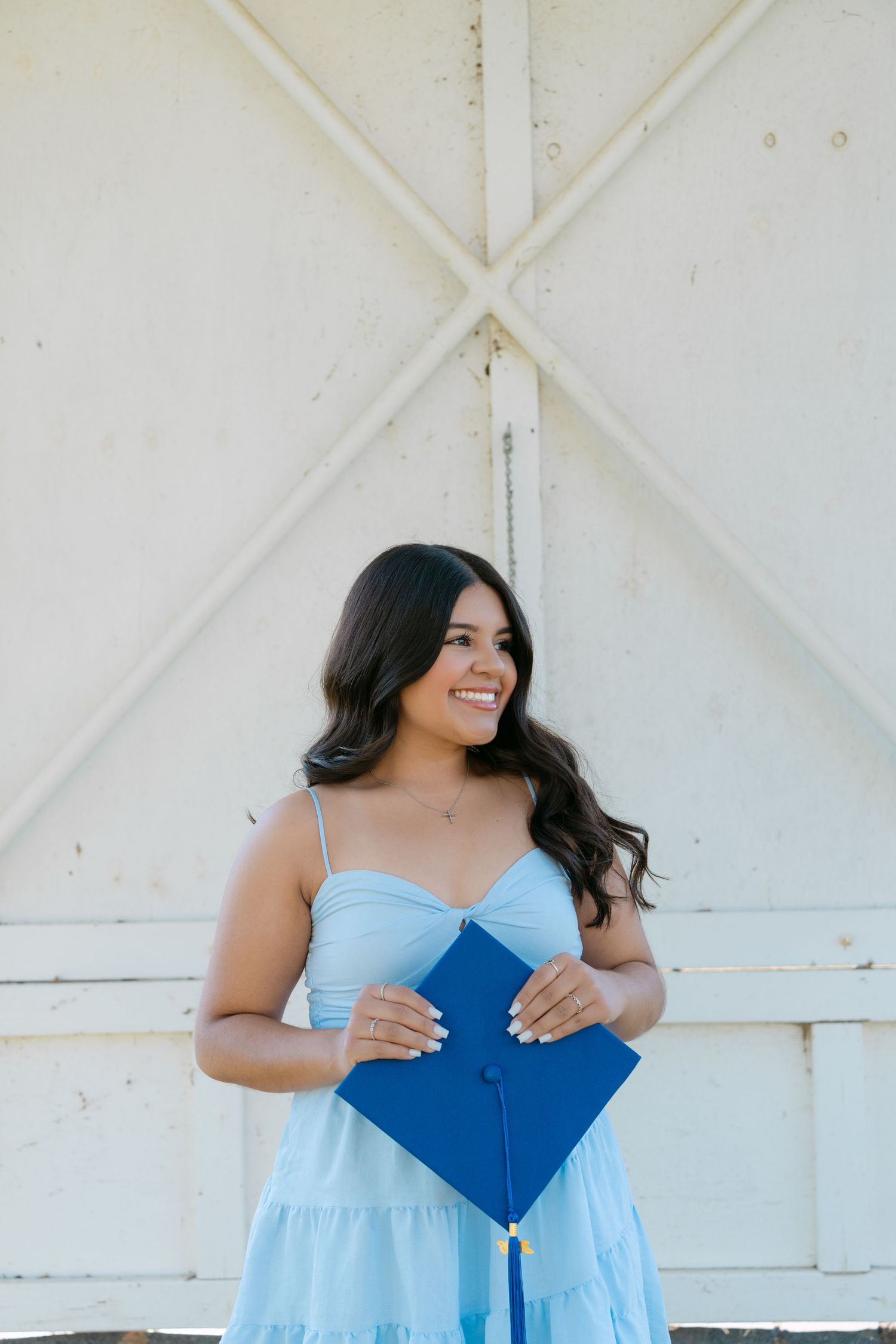 Smiling young woman in light blue dress holding blue graduation cap outdoors against white barn door.