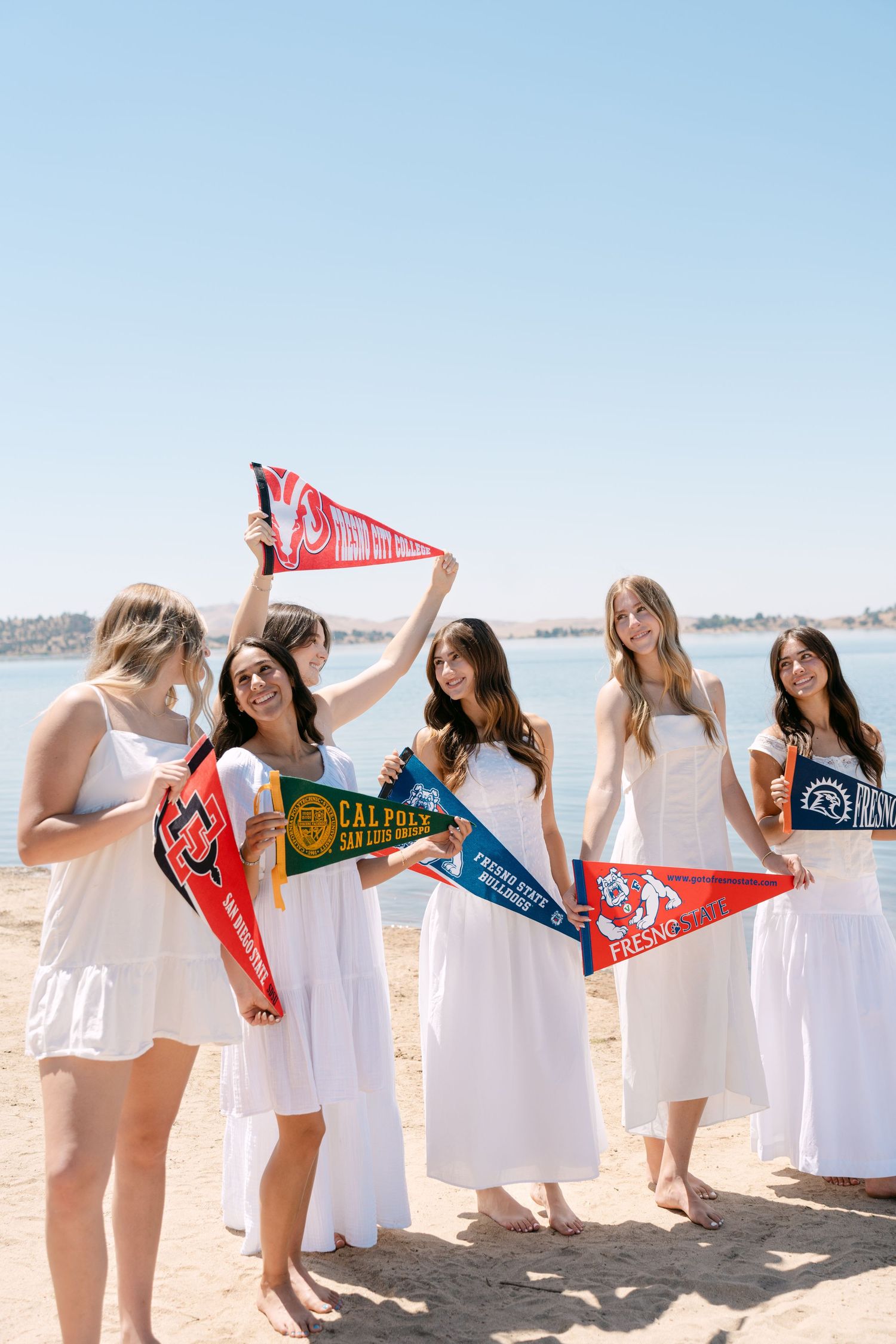 Group of young women in white dresses holding college pennants on a sunny beach near calm blue water.