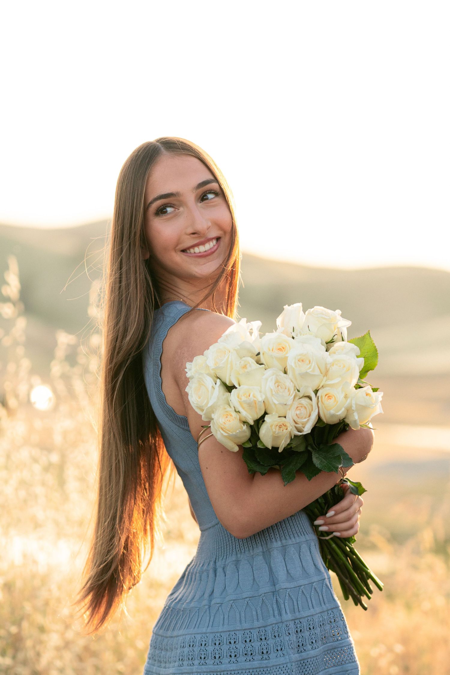 A woman in blue dress holds a bouquet of white roses during golden hour.