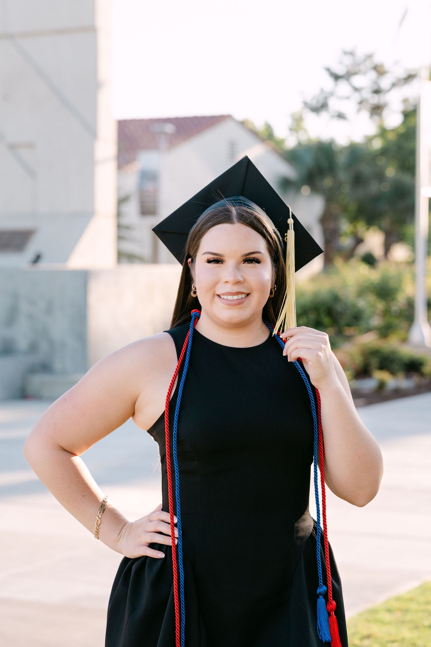 Smiling graduate in black cap and gown with red and blue stoles poses outdoors on campus.