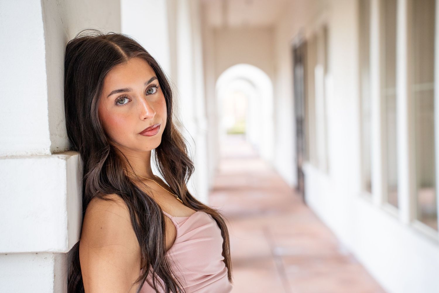 A woman in a pink top poses against a white archway in an elegant corridor.