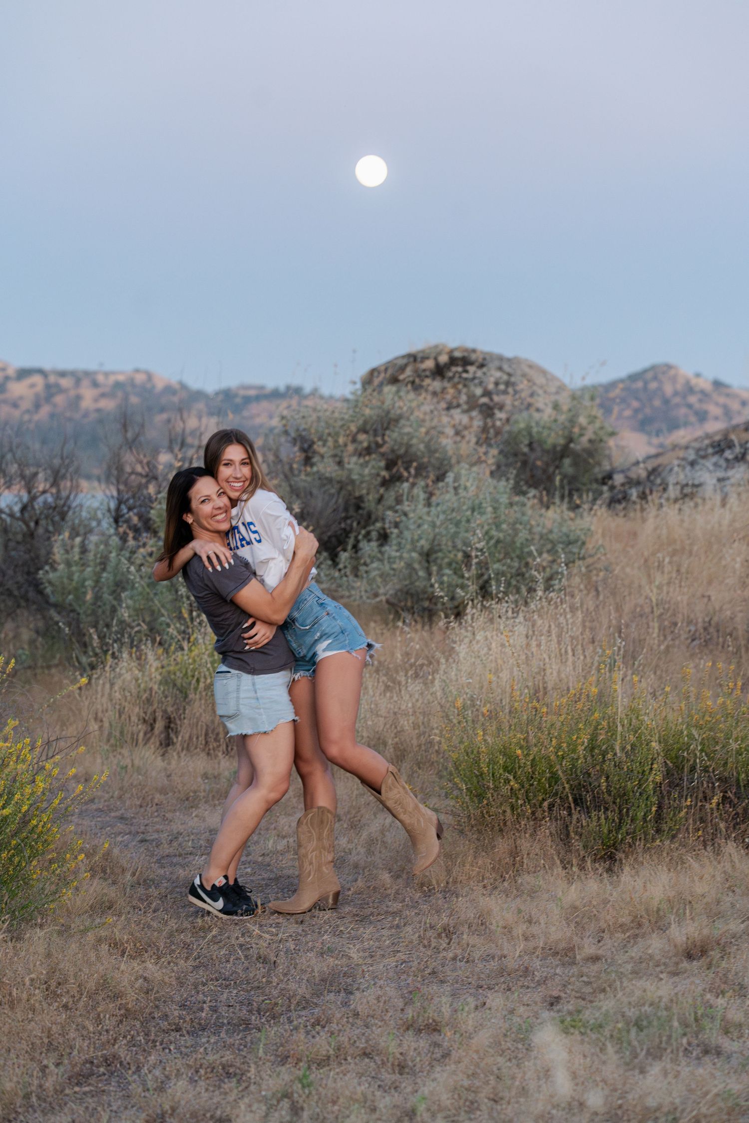 Two people embrace and laugh together under a full moon in a mountain landscape.
