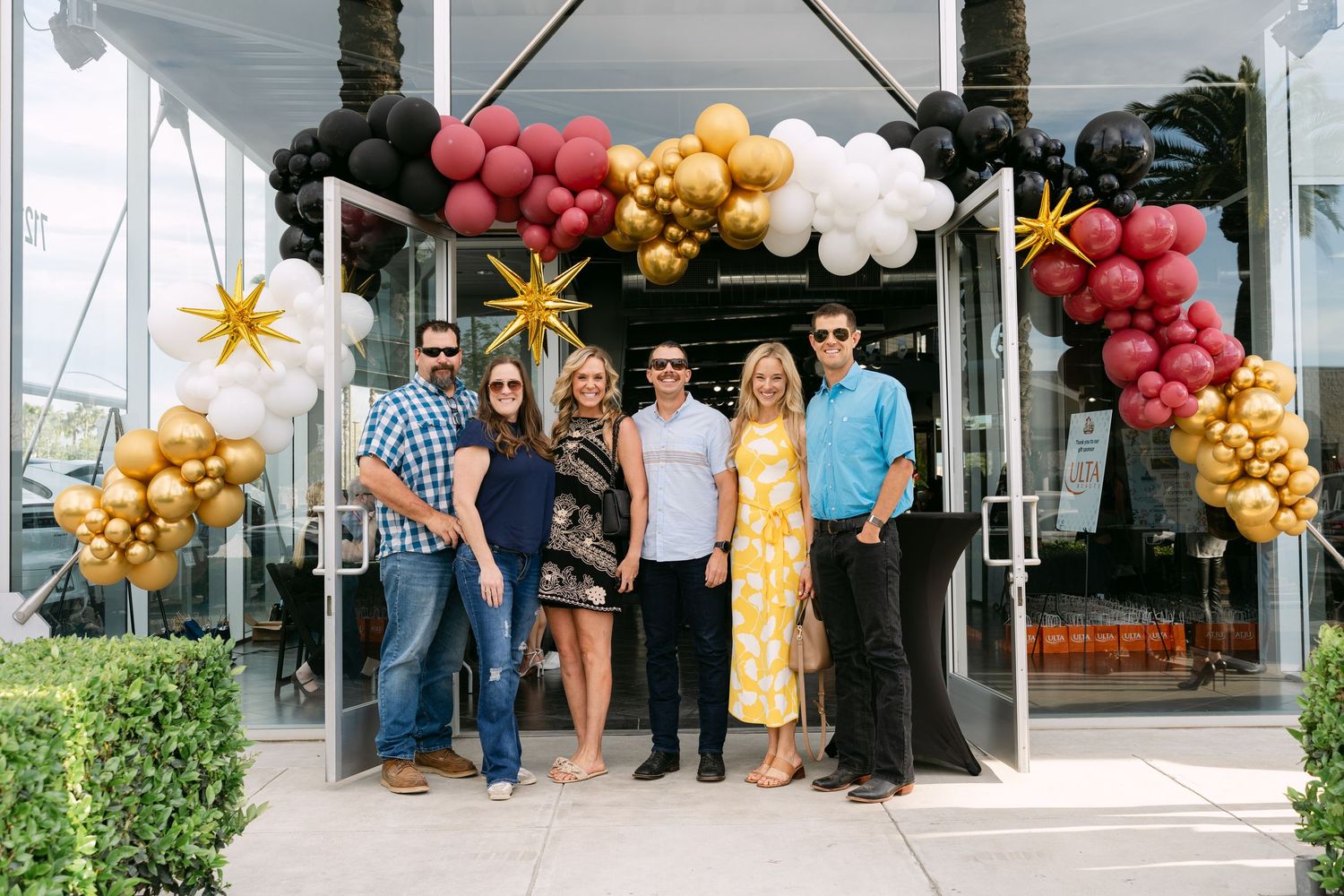 A group of people stand outside a building entrance decorated with pink, gold, and black balloon arches and star decorations.