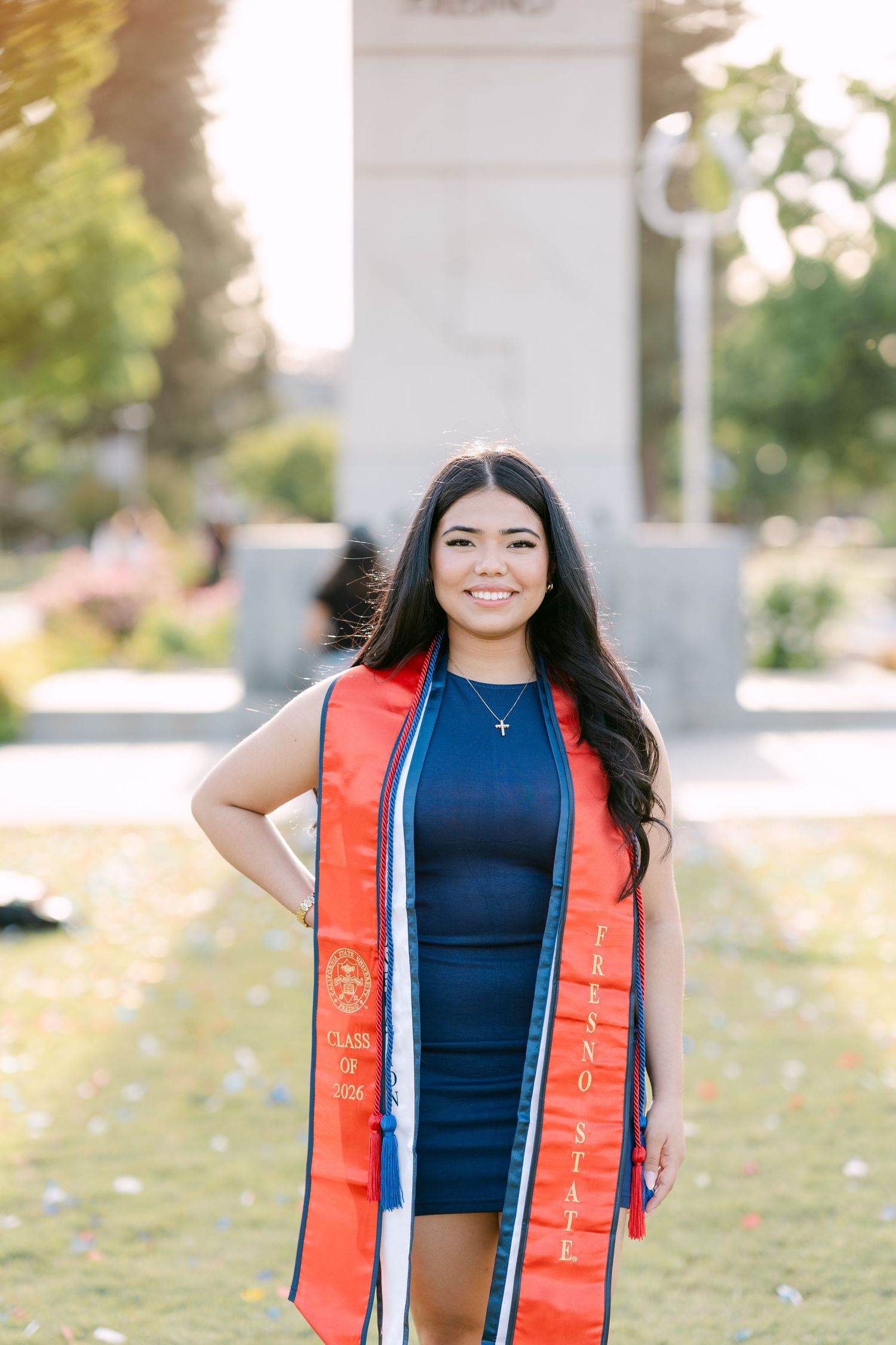 Smiling college graduate in navy dress and orange Fresno State stole poses outdoors on sunny campus.