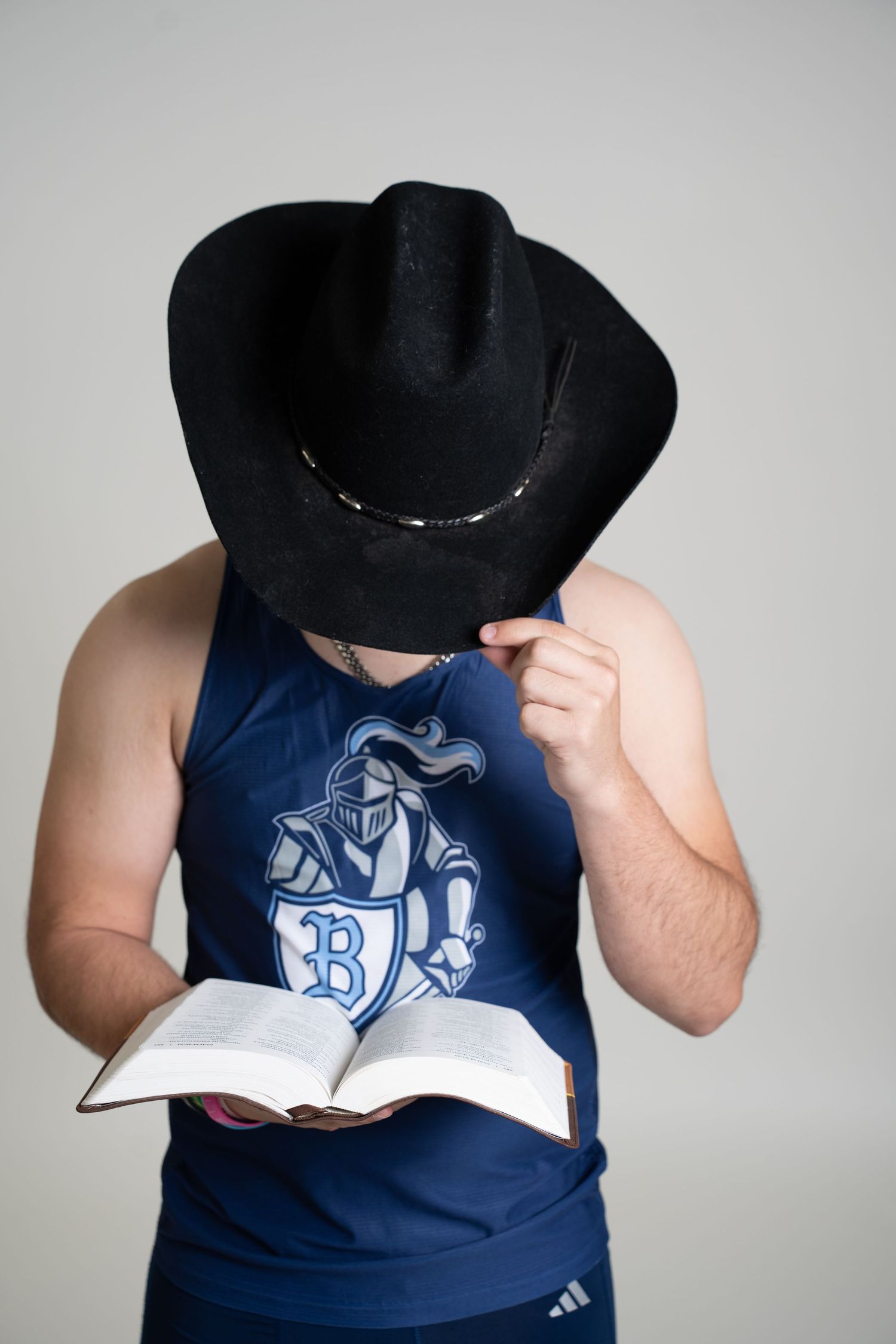 Person in a blue tank top and black cowboy hat reads a book while looking downward.