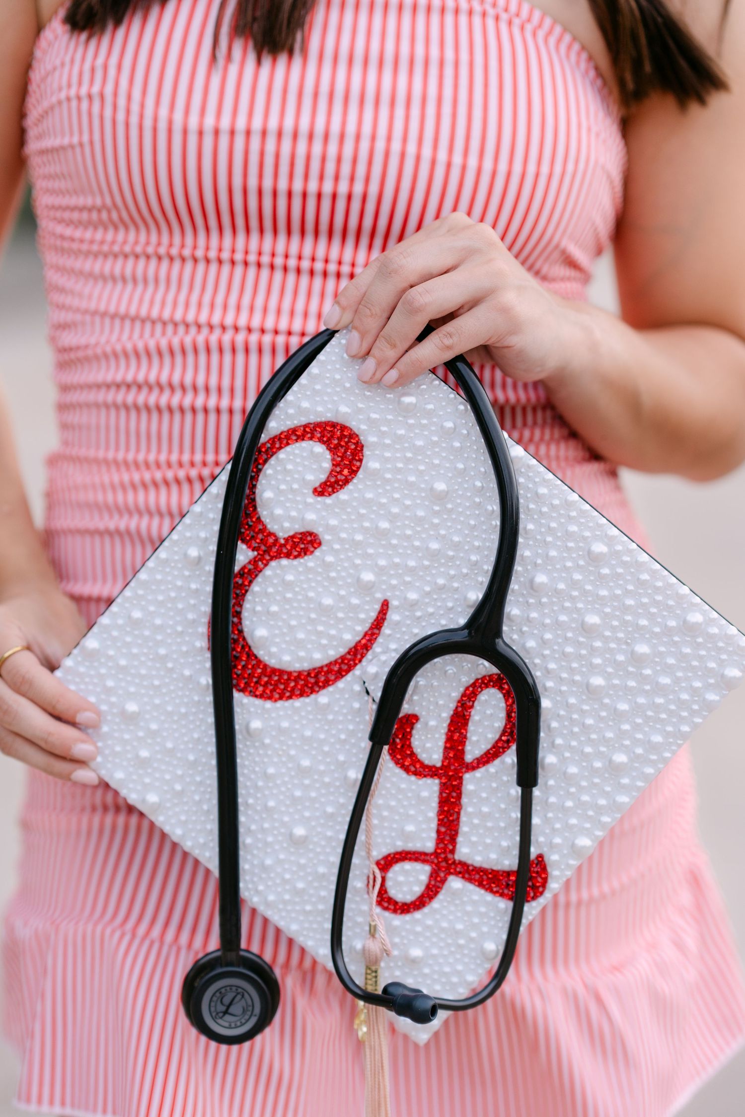 Decorated graduation cap with red rhinestone initials E and L, adorned with pearls and a black stethoscope.