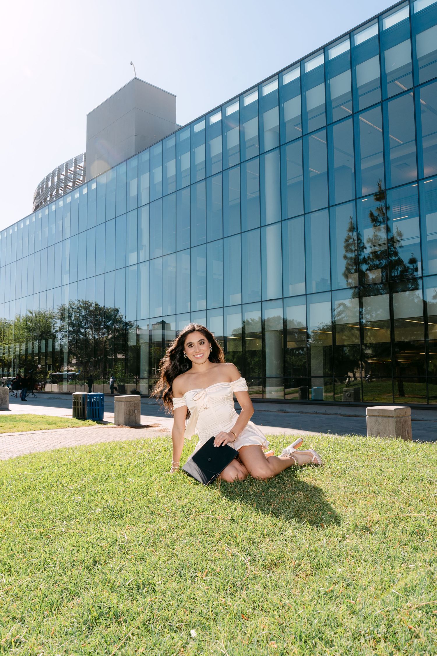 Young woman in off-shoulder pink top sits on grass in front of modern glass building on sunny day.