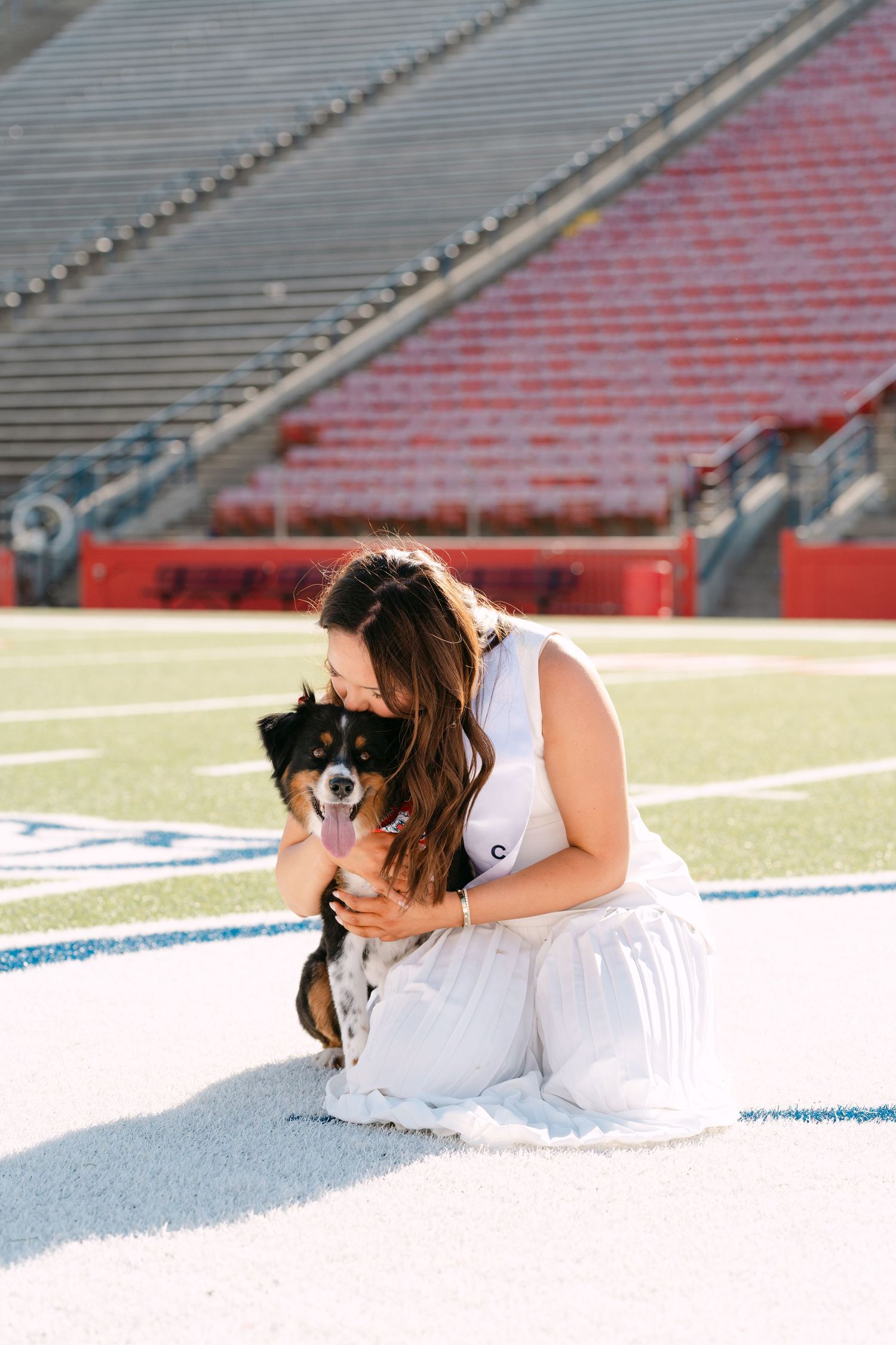 Woman in white dress kneeling on football field turf, kissing small black and tan dog in empty stadium.