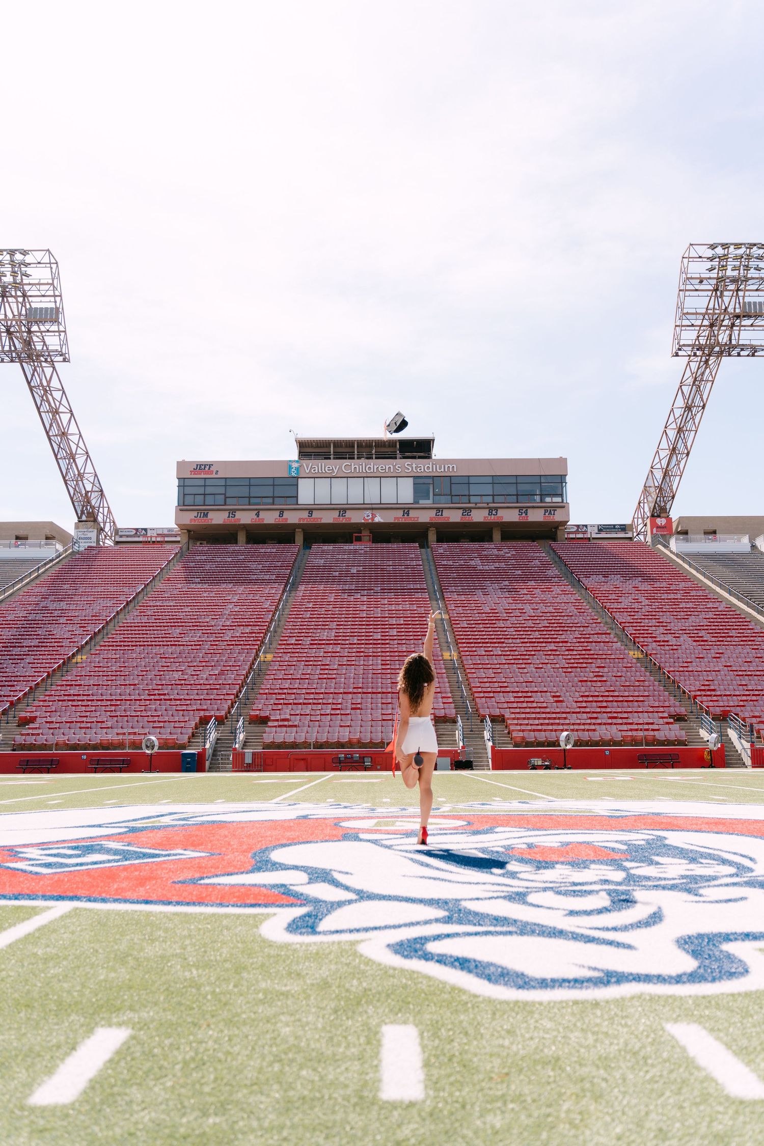 Woman standing on football field at empty Fresno State Bulldog Stadium with red bleachers and scoreboard.