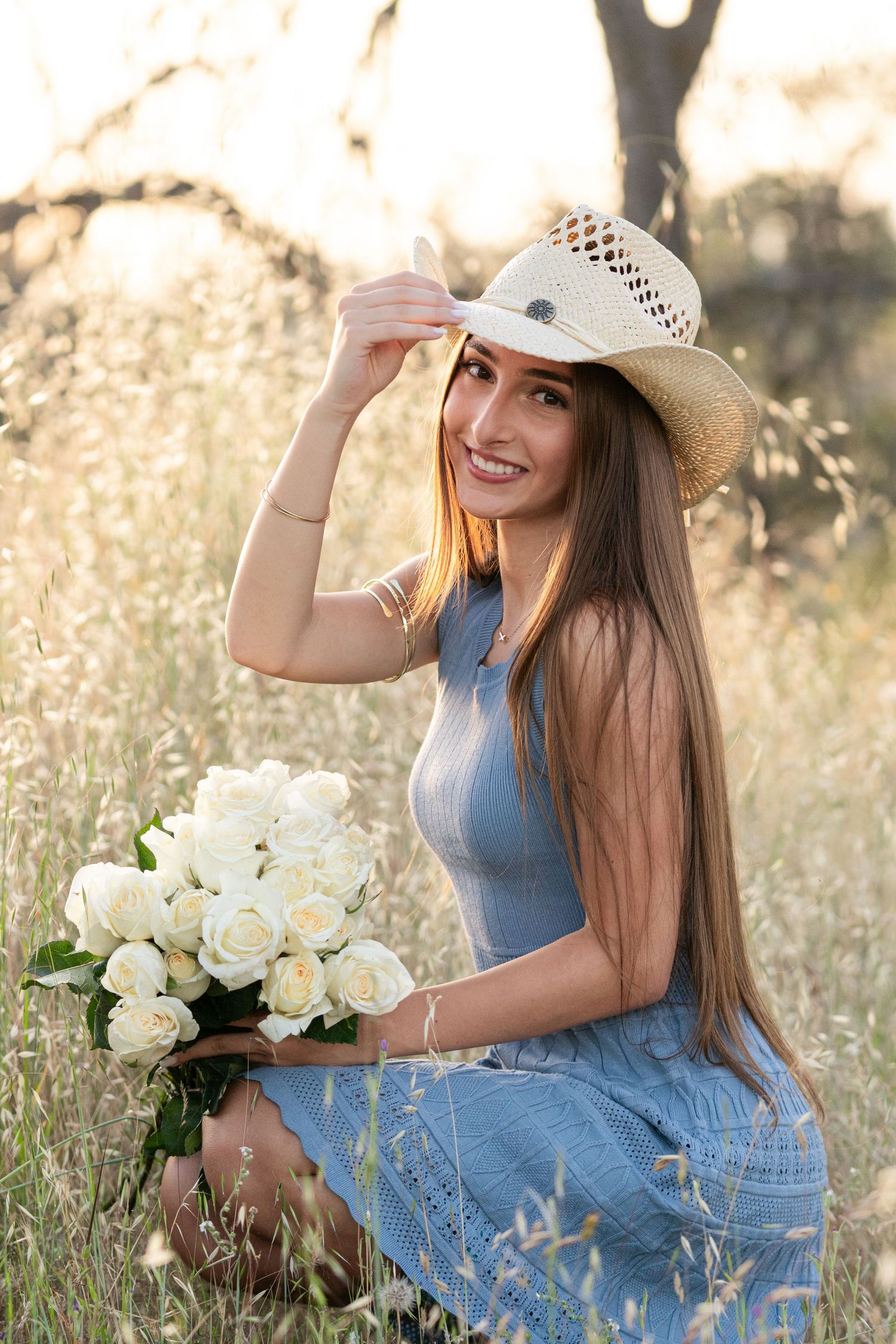 Young woman in blue dress and straw hat holding white roses bouquet in golden field at sunset.
