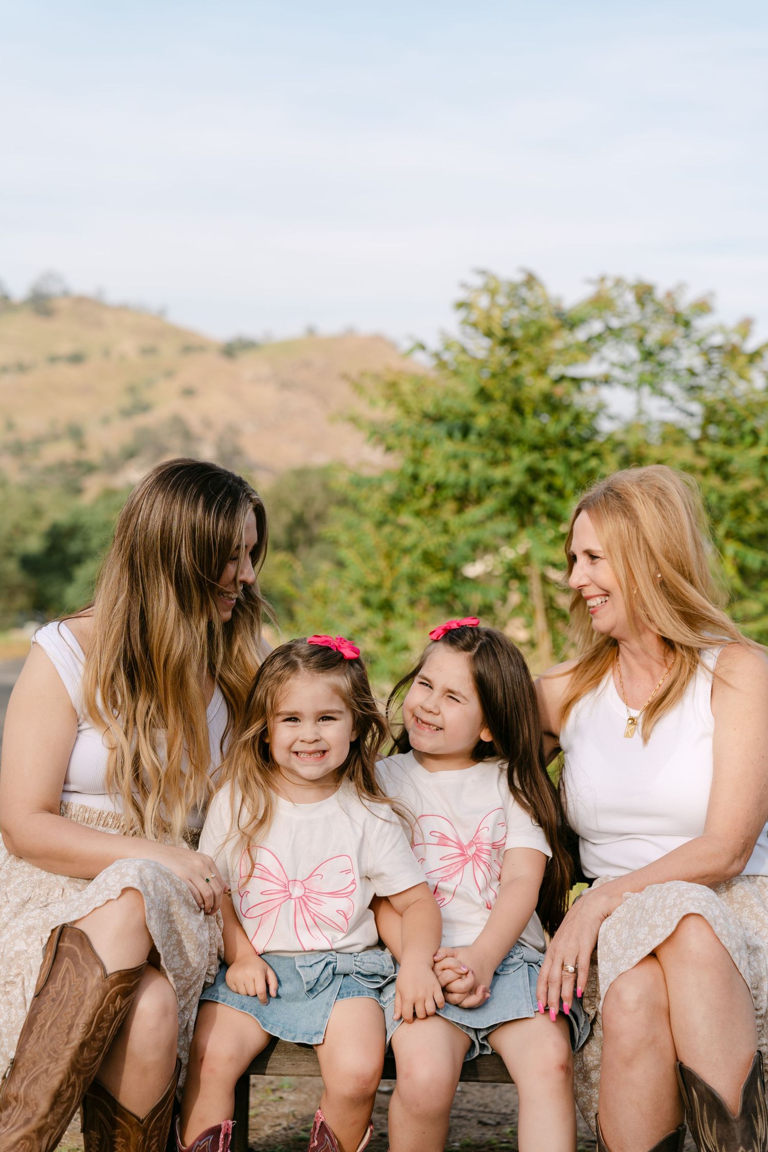 Two women and two little girls in matching bow shirts sit together on a bench outdoors with green hills behind them.