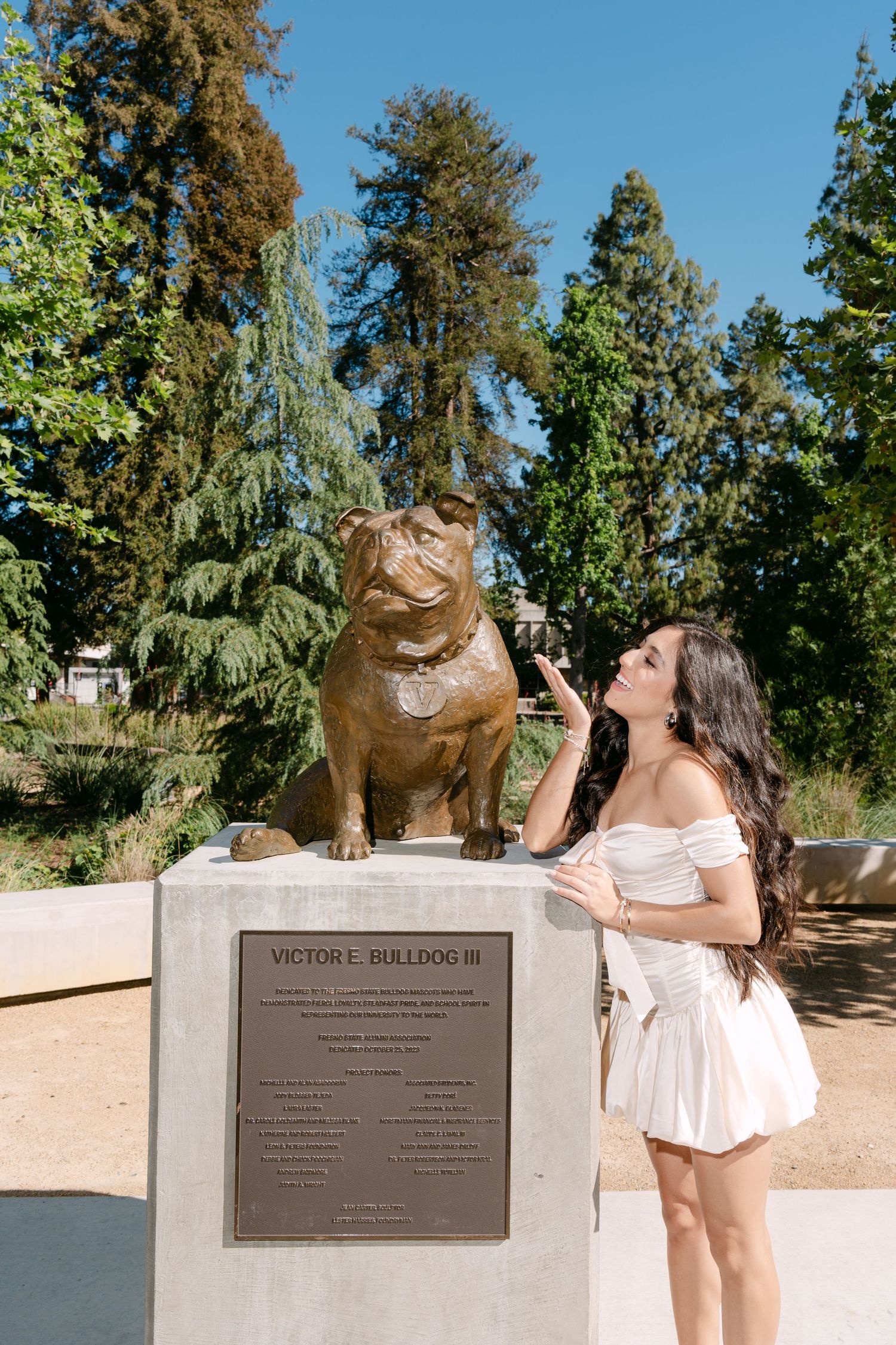Young woman in white dress posing with Victor E. Bulldog III bronze statue on sunny campus day.
