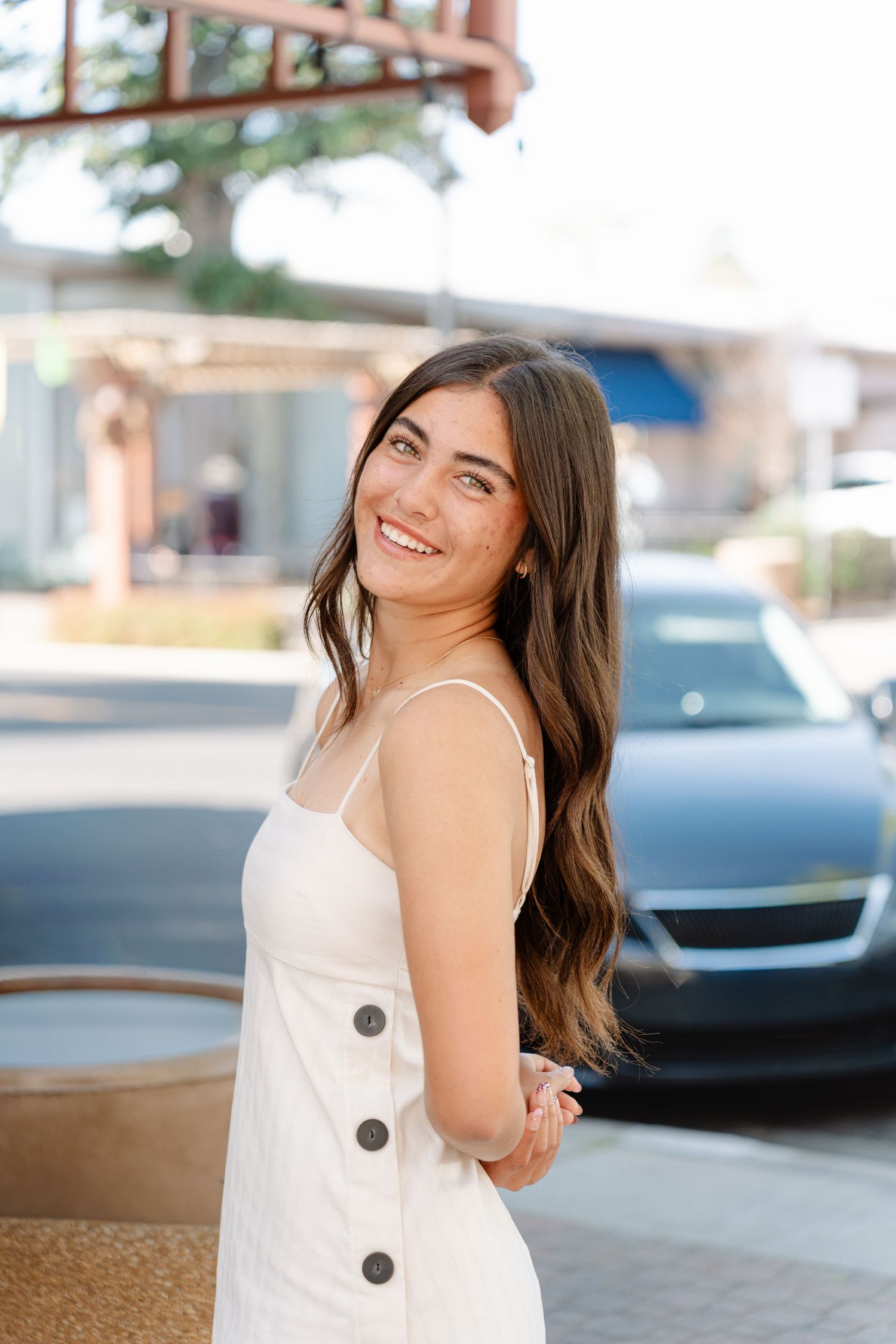 A person in a white sleeveless top with decorative buttons poses outdoors near parked cars.