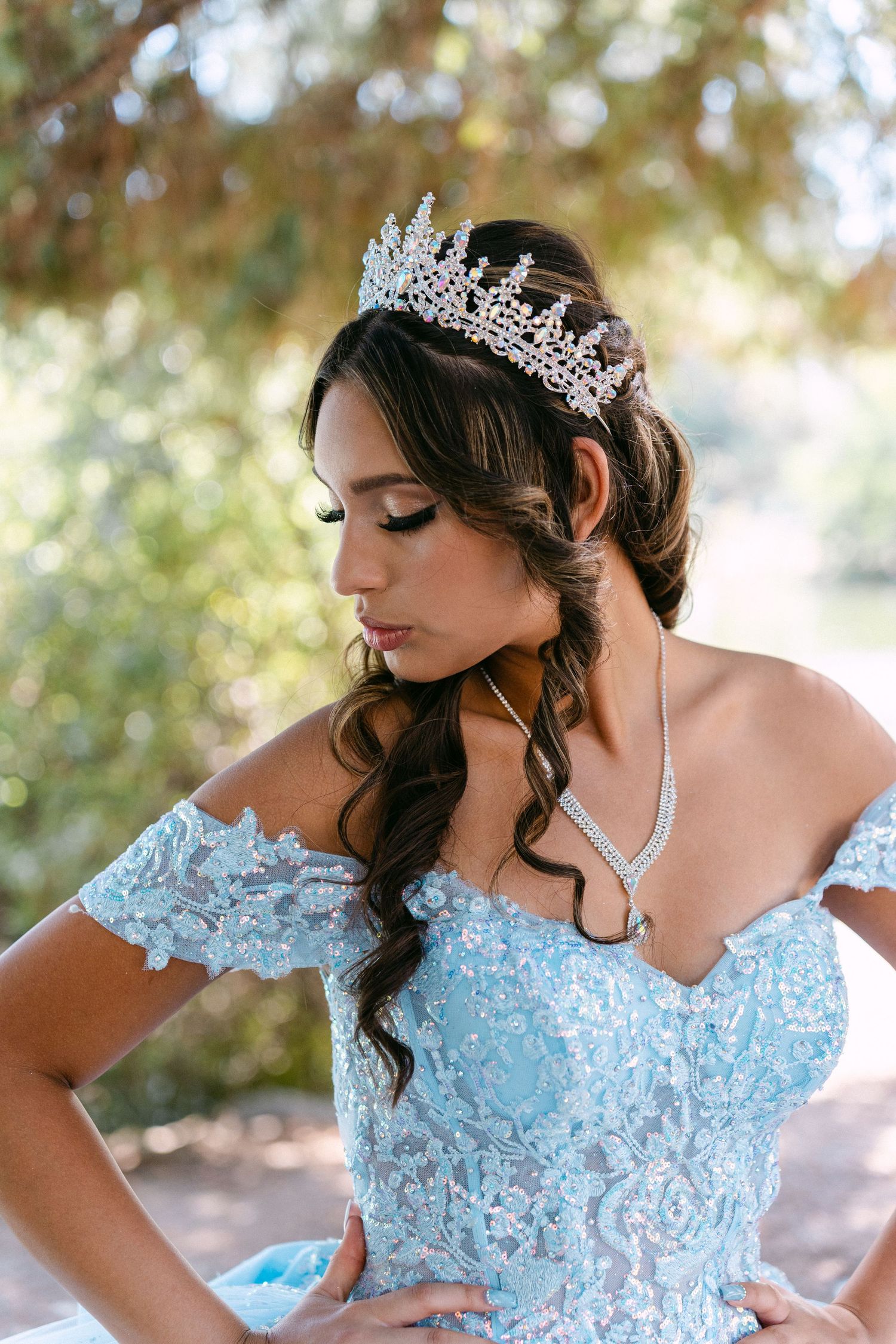 A person wearing a light blue formal gown and tiara poses in an outdoor setting.
