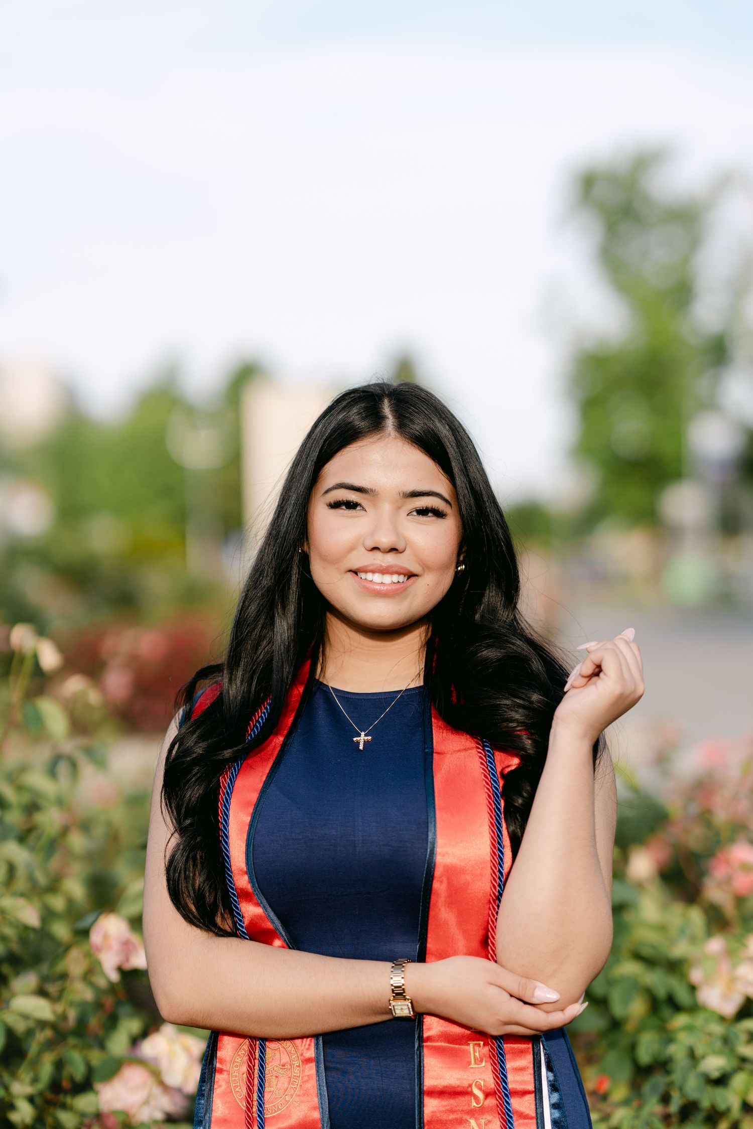 Young woman in navy blue graduation attire with red sash smiles confidently outdoors among blooming roses.