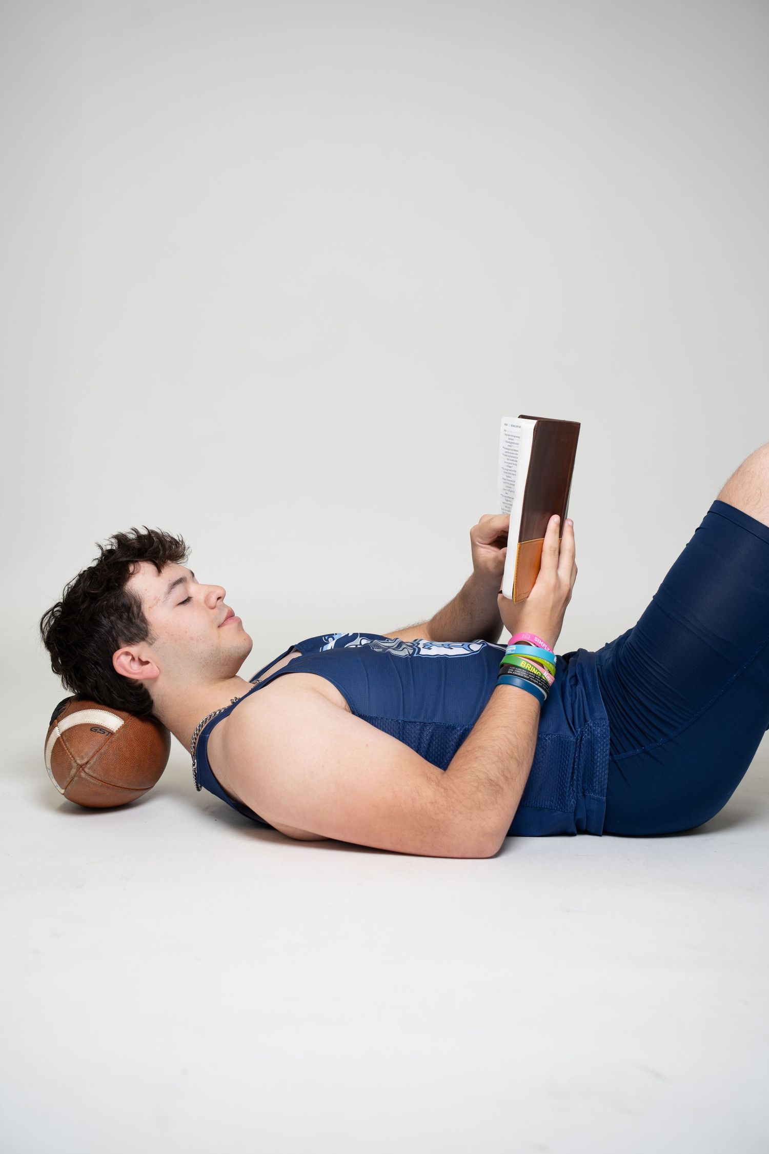Person lying on floor reading book with football as pillow while wearing navy blue athletic outfit.
