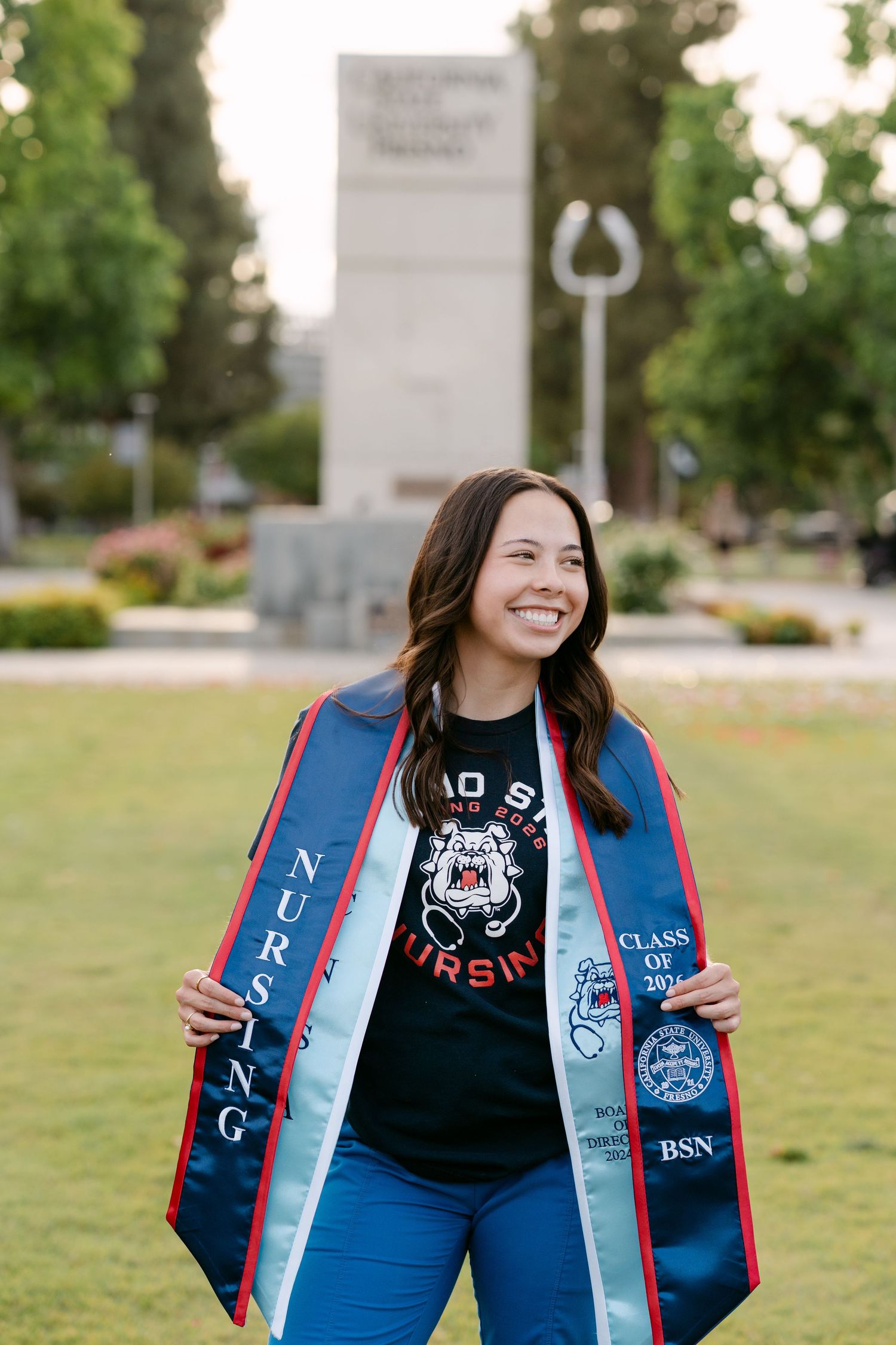 Smiling nursing graduate proudly displaying her Fresno State BSN graduation stole outdoors on campus.
