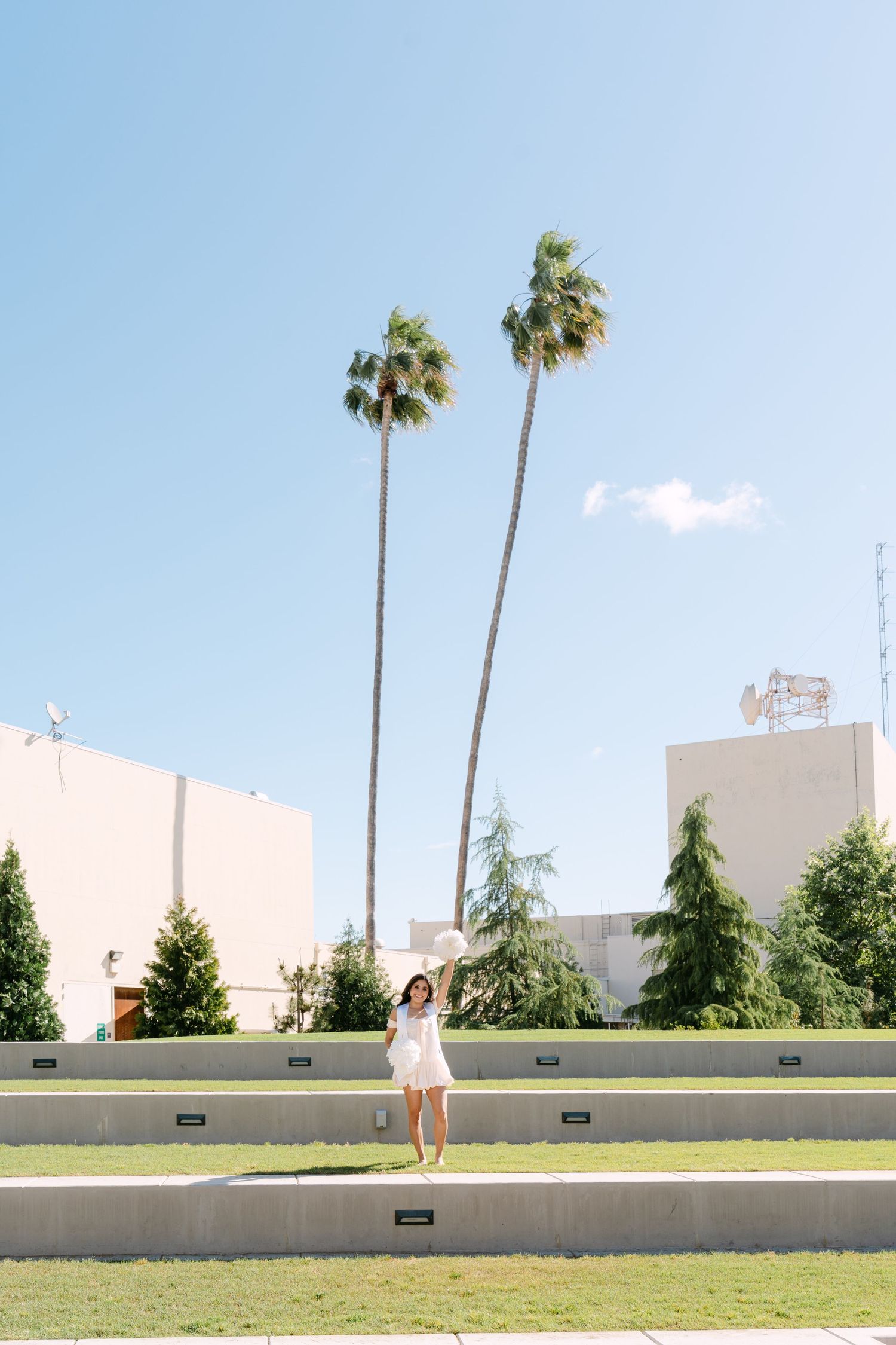 Woman in white dress standing on steps with tall palm trees and blue sky in an urban plaza setting.