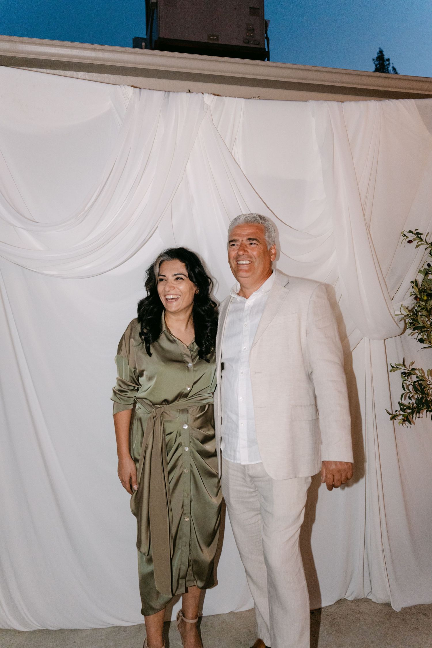 A couple poses together at an event, with the woman wearing an olive green dress and the man in white attire.